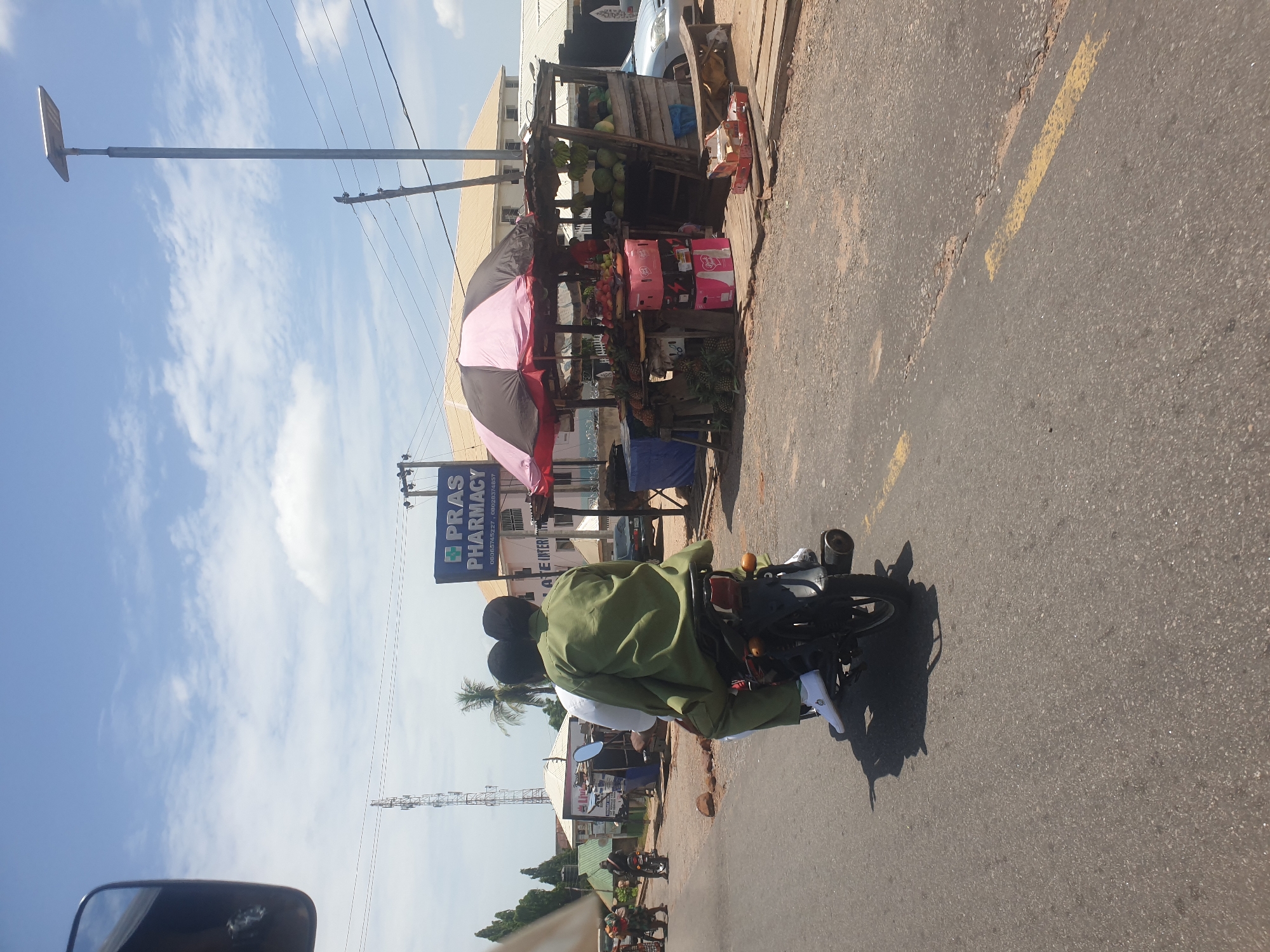 A street scene in Zango-ndaji, Nigeria, shows a motorcycle with two occupants traveling on an asphalt road with a yellow centerline. The passenger is wearing a green uniform with white shoes. A fruit stand is set up on the side of the road, shaded by a large pink and black umbrella. In the background, a sign for "Pras Pharmacy" is visible. Utility poles and a communication tower are visible in the background. Additional people and motorcycles are further down the road. The sky is partly cloudy.