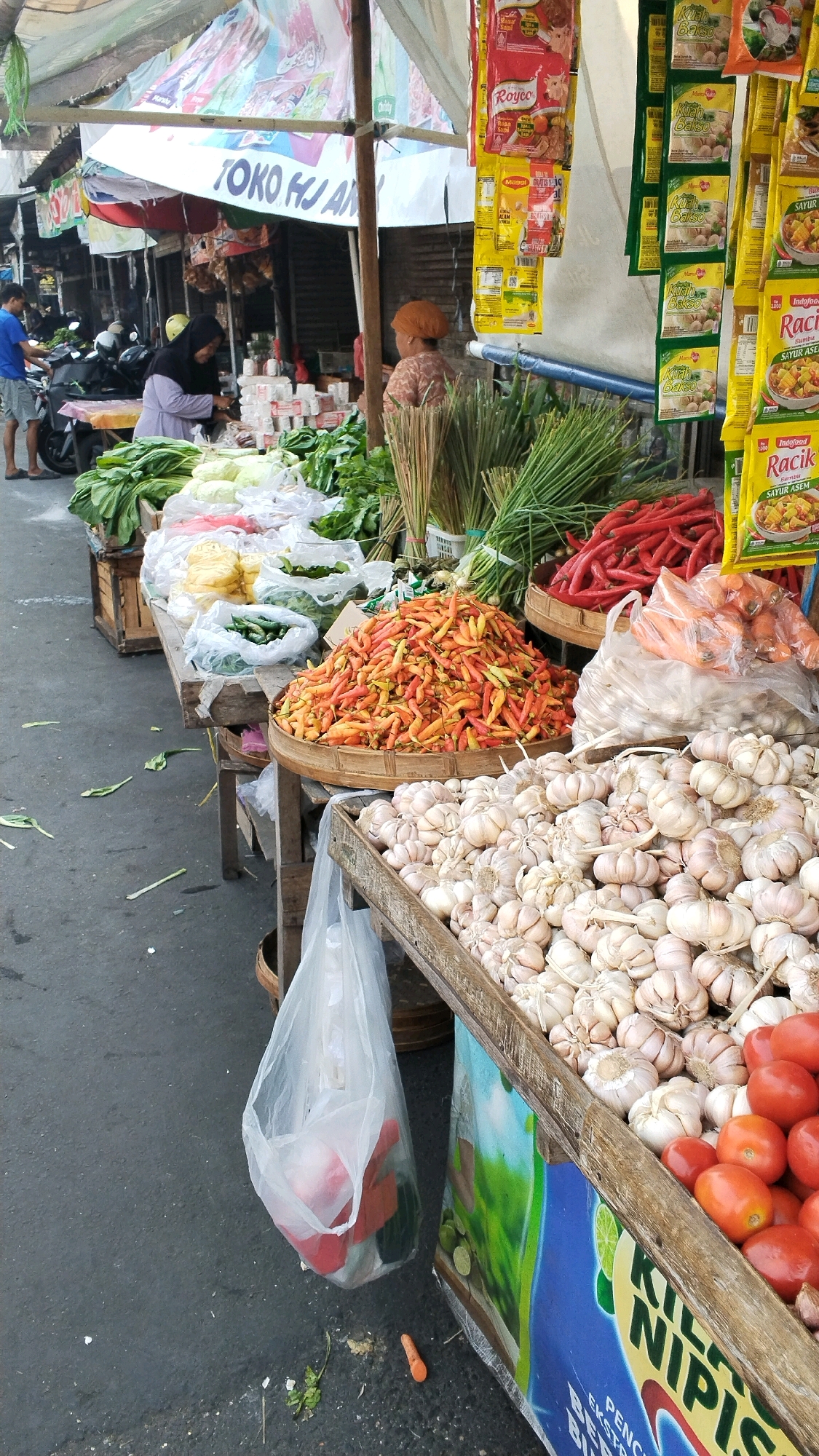 This is a medium shot of an outdoor market stall in Mojokerto, Indonesia. The stall is laden with fresh produce and packaged goods. In the foreground, a wooden crate overflows with bulbs of garlic, and to the right, a wooden bin is filled with ripe red tomatoes. Behind the garlic and tomatoes, a large pile of small, multi-colored chilies sits in a woven bamboo basket. Further back, bundles of fresh green onions and red chilies are visible. Various green leafy vegetables are displayed in plastic bags and wooden crates. Packaged food items, including small packets of seasonings and crackers, hang from the stall's canopy.
Two individuals are present. A woman wearing a purple hijab and light-colored clothing is standing behind the stall, appearing to be interacting with the produce. Another person, wearing an orange headscarf, stands to the right, facing away from the camera.
The market appears to be bustling, with a motorcycle parked behind the stall, suggesting a busy atmosphere. The ground is paved and somewhat weathered, with a few stray leaves scattered on it. The overall impression is of a vibrant, local market scene.