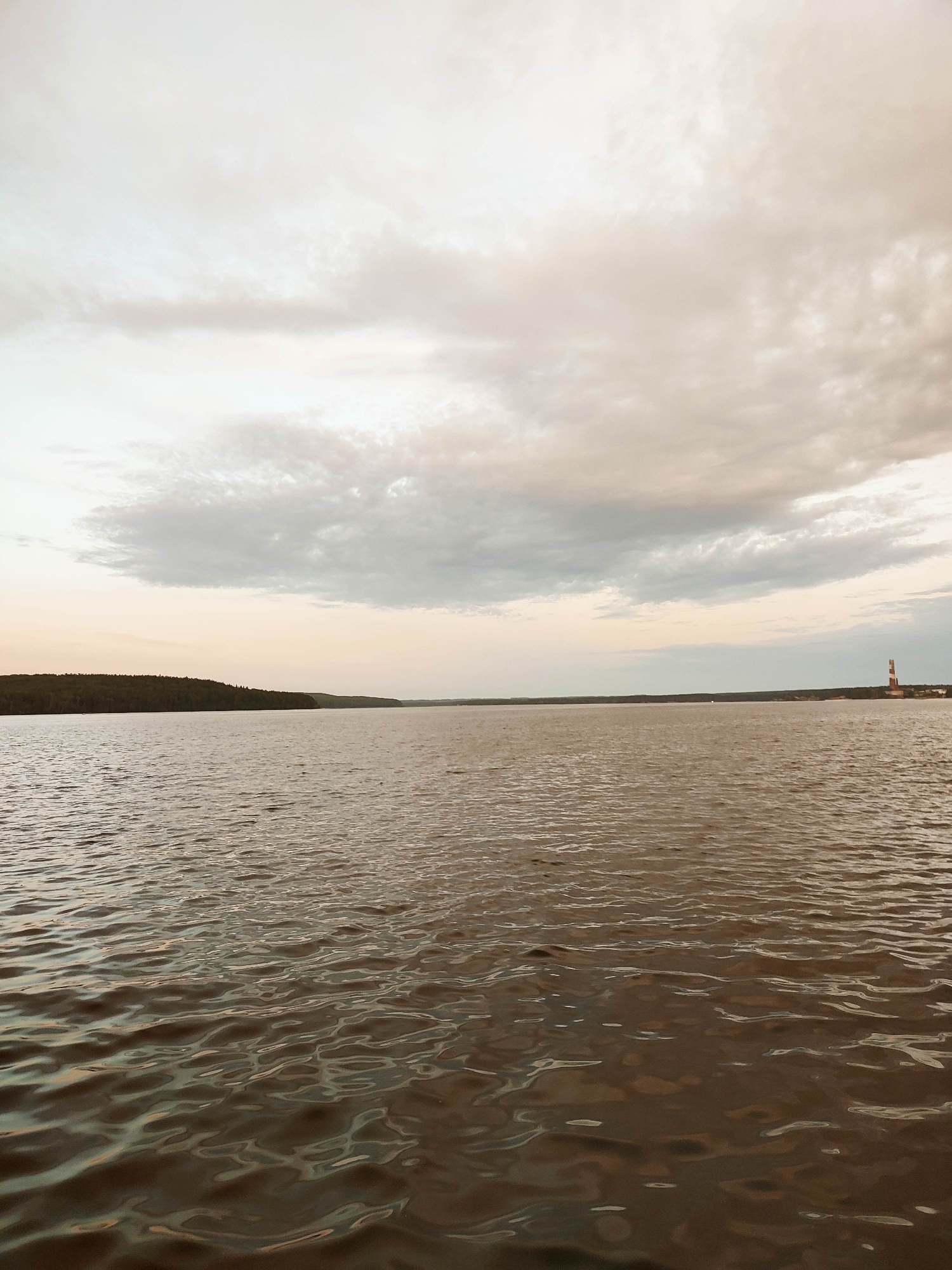 The image depicts a wide body of water under a cloudy sky. The water is a murky brown color, with visible ripples and reflections. In the distance, a tree-covered shoreline stretches across the horizon. To the right of the frame, a red and white striped lighthouse stands near the shore. The lighting suggests it is either dawn or dusk, with the sky being a pale gray and pinkish hue. There are no people or animals visible in the image.