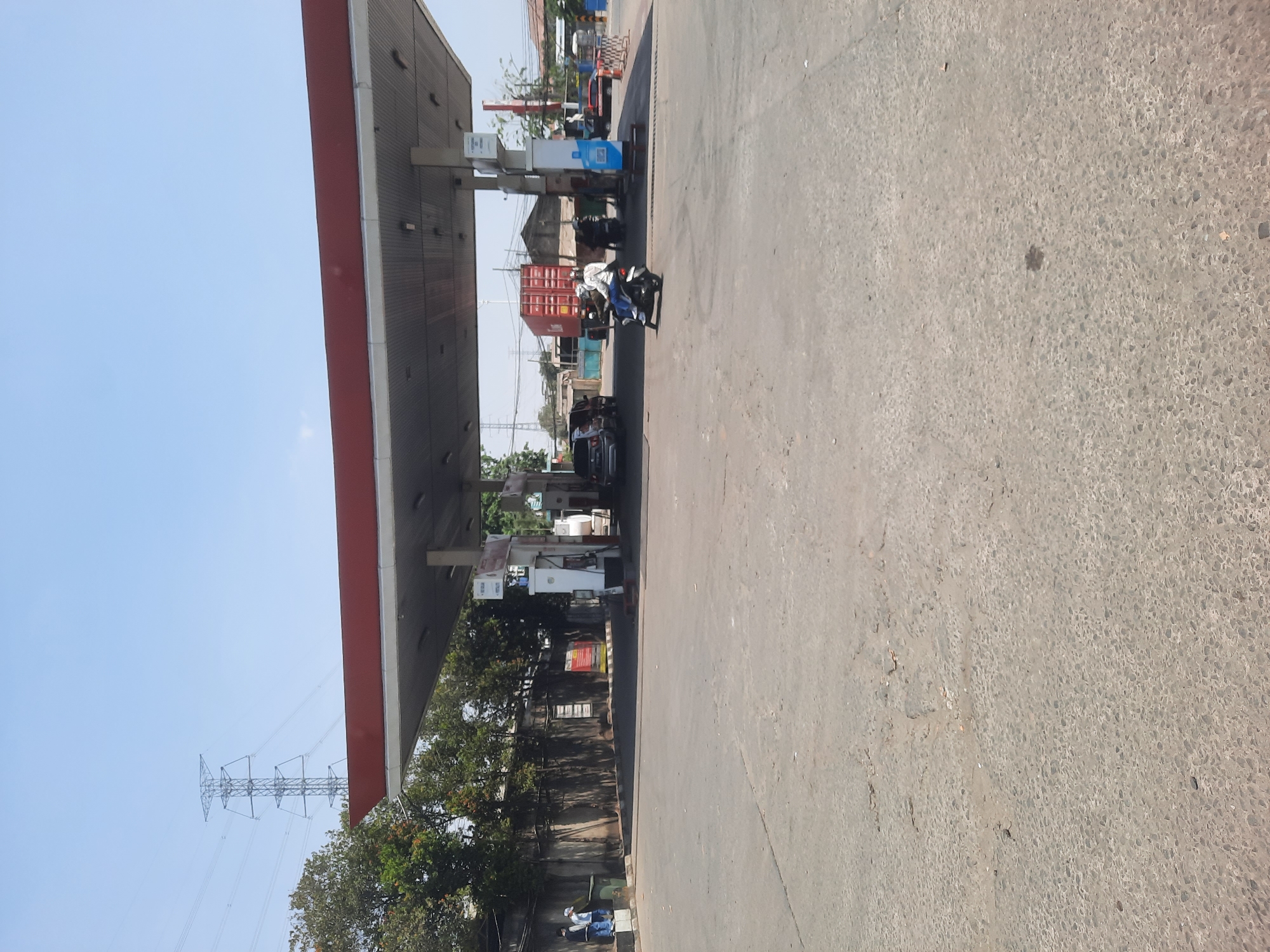 A street-level view depicts a gas station with a red-roofed canopy, supported by white pillars. Various vehicles are present, including a motorcycle with a rider wearing a white jacket and blue jeans, a black SUV, and another motorcycle. In the background, a large red shipping container is visible on a truck. The ground is paved with asphalt and appears weathered with some cracks. Trees and a tall transmission tower are seen in the distance, against a clear blue sky. The scene is likely in North Jakarta, Indonesia, given the visual cues of urban infrastructure and vegetation.