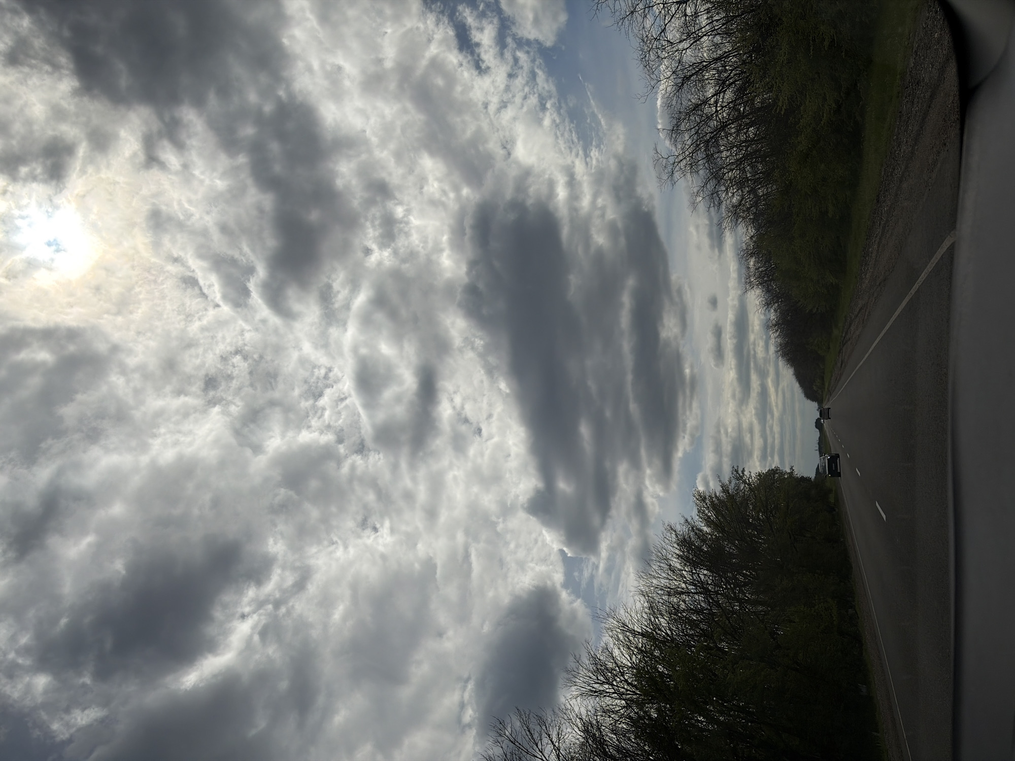 A photograph taken from inside a vehicle shows a grey asphalt road flanked by dense trees on either side. The sky overhead is filled with a variety of grey and white clouds, with the sun faintly visible through the cloud cover on the left side of the image. The road leads into the distance, where a few vehicles can be seen. The image appears to be taken while traveling on a road outside Starominskaya, Russia.
