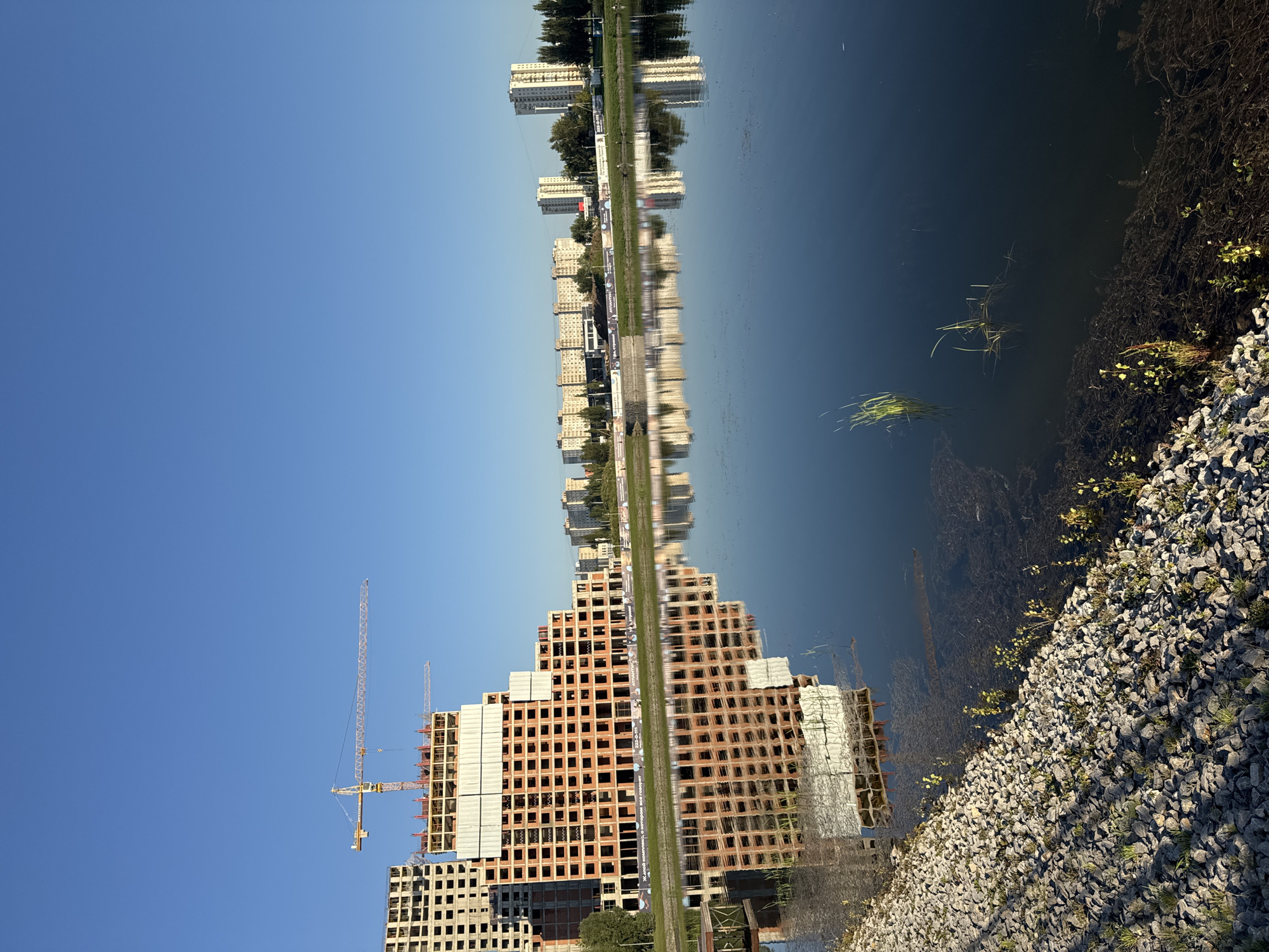 The image depicts a construction site of multi-story residential buildings with a lake in the foreground. The buildings are in various stages of construction, with the primary focus being a large, unfinished orange brick structure with exposed concrete floors and columns. A tall yellow construction crane is visible above this structure. In the background, several completed apartment buildings and other structures are visible, suggesting an urban or suburban setting. The lake reflects the sky and the buildings, creating a mirror-like effect. The shore of the lake is lined with grey gravel and some sparse vegetation. The sky is clear and blue, indicating a sunny day. The scene appears to be located near the "Atlantic City" residential complex in Ufa, Russia, as indicated by text on a banner visible in the mid-ground. There are no people visible in the image.