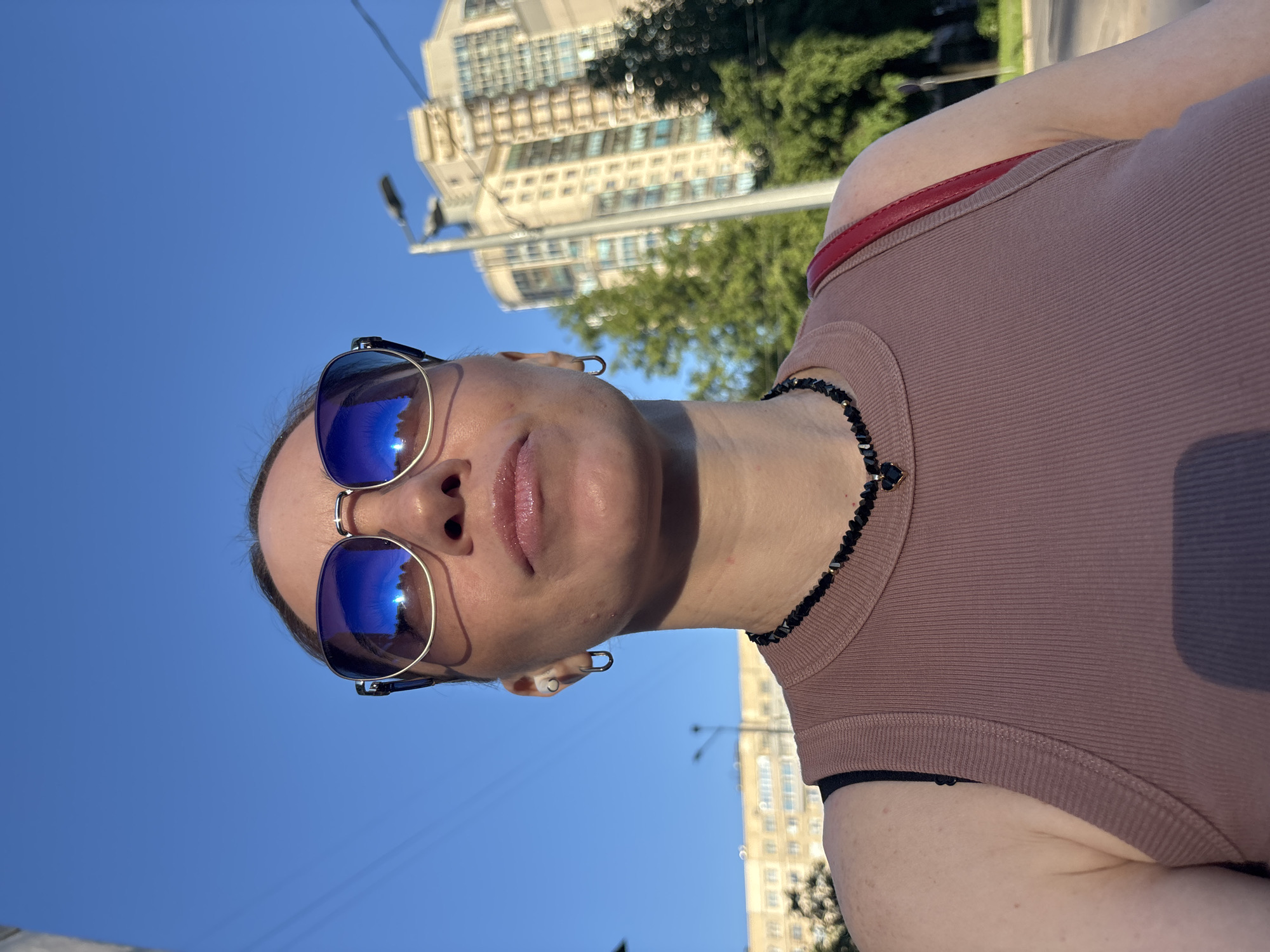 The image presents a low-angle, upward-facing shot of a woman in an urban setting within the city of Sinyavino, Russia, under a clear blue sky. The woman is visible from the chest up, wearing blue reflective aviator-style sunglasses with gold-toned frames that obscure her eyes. Her mouth is closed, with visible glossy lips. She has a white earbud in her left ear and a gold-toned hoop earring in her right ear. Around her neck is a black beaded necklace featuring a small black heart-shaped pendant with gold accents. She is dressed in a light brown, ribbed tank top; a black bra strap is visible on her left shoulder. A red strap, consistent with a shoulder bag, passes over her right shoulder. The background, out of focus, includes several multi-story buildings, notably a taller, light-colored building with multiple windows on the right side of the frame. Green foliage from trees is visible between and in front of the buildings. A street light pole extends vertically on the right side, and thin wires crisscross the expanse of the bright blue sky. The scene is brightly lit by natural sunlight.