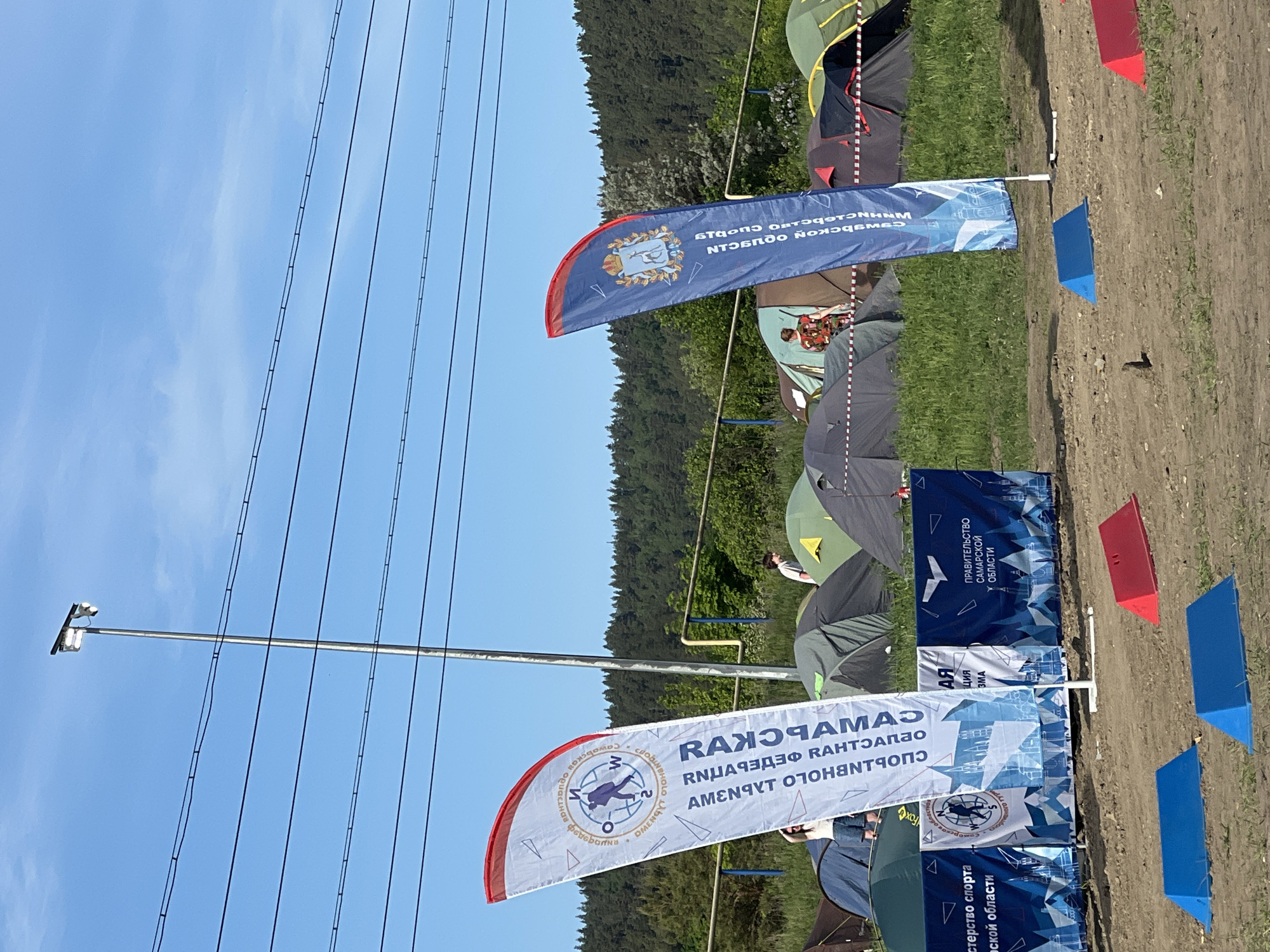 The image shows an outdoor event, possibly a sports competition or festival, taking place in Khvalynsk, Russia. Several tents are set up on a grassy area, and a series of flags line a path or boundary. The flags display branding and logos, including those of the Samara region government, the Ministry of Sports of the Samara region, and an event titled "RANOMAMAZ". Blue and red markers are placed alongside the flags. Overhead power lines are visible against a clear blue sky, and a forested area forms the background. People are visible inside some of the tents.