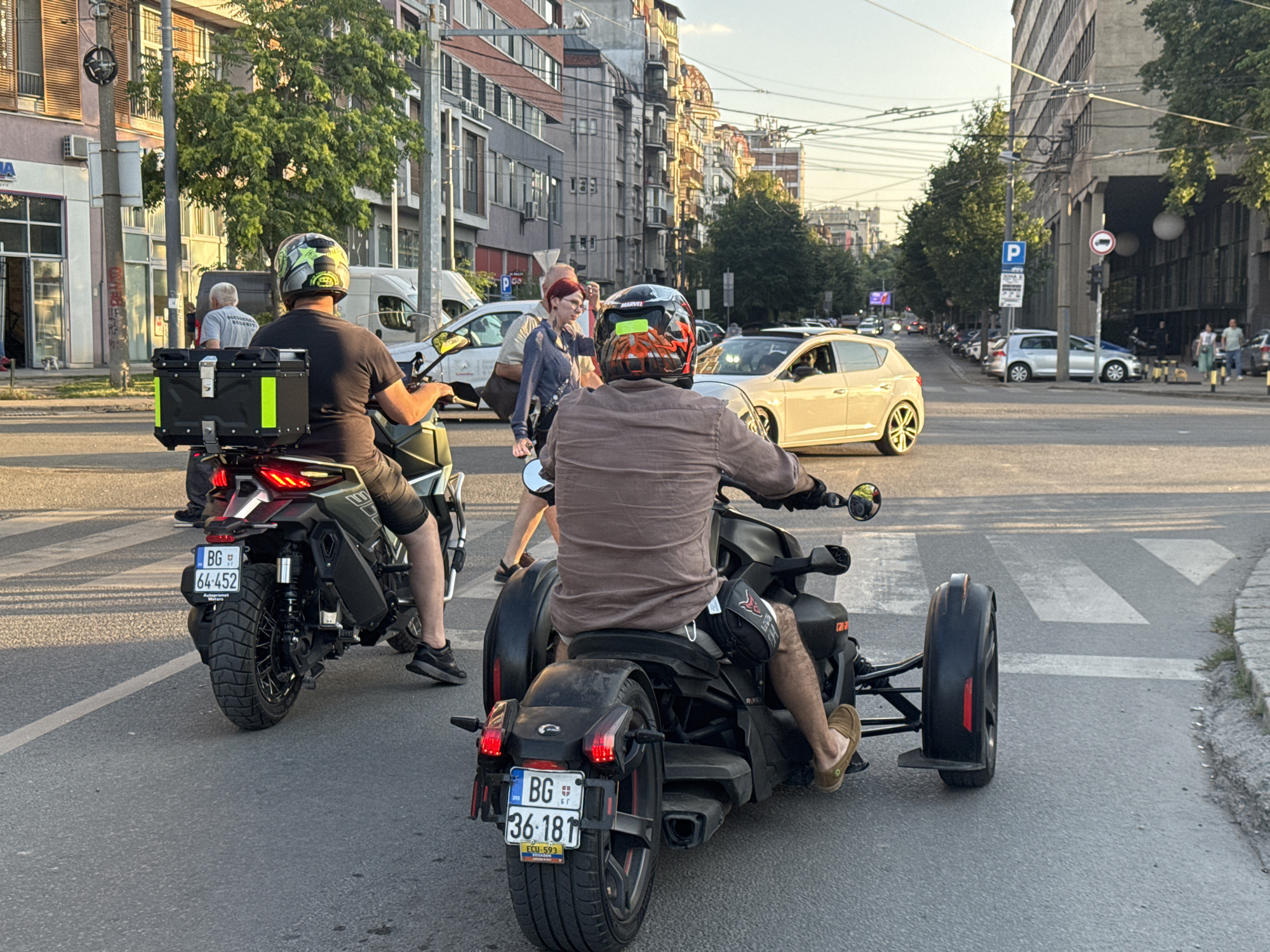 The image captures a street scene in Belgrade, Serbia, during what appears to be late afternoon or early evening. The perspective is from behind two three-wheeled vehicles, likely Can-Am Rykers, moving on a paved road. The vehicle in the foreground has a license plate that reads "BG 36-181". The rider is wearing a brown shirt, shorts, and brown slip-on shoes, with a black bag slung over their thigh. The vehicle behind it has a license plate that reads "BG 64-452". Its rider wears a black t-shirt, jeans, and black athletic shoes, and a black storage box is mounted on the rear.
The environment is urban, characterized by multi-story buildings with visible architectural details, including balconies and windows, lining both sides of the street. Trees with green foliage are interspersed along the sidewalks. There are other vehicles visible, including a white sedan in the middle ground and several cars parked along the right side of the street. Pedestrians are also present, with a few individuals walking on the sidewalk in the background. Overhead, electrical wires crisscross the sky, indicating trolleybus or tram lines. The lighting suggests the sun is low, casting long shadows. A pedestrian crossing is marked on the road in the foreground.