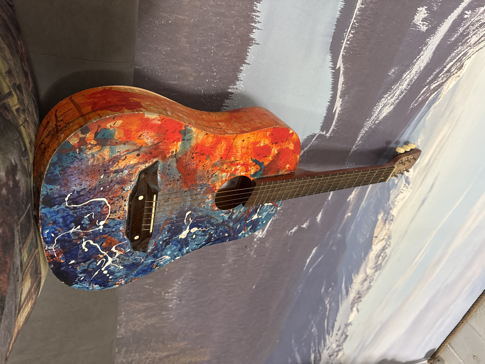 An acoustic guitar with a unique paint job is positioned in front of a wall with a blurred mountain scene. The guitar's body is painted with a combination of blue, orange, red, and white paint splatters. It appears to be leaning slightly against the wall. The background features shades of blue, gray, and white, resembling a mountain landscape. The image appears to have been taken at a slightly oblique angle, and the location, based on the prompt, is Chaikovsky, Russia.