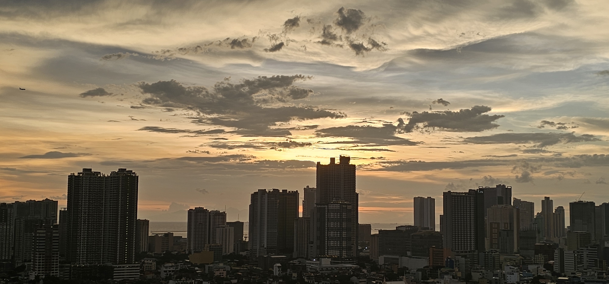 A wide-angle, high-angle shot captures a cityscape at sunset in Makati, Philippines. The sky is filled with dramatic clouds, painted in hues of orange, yellow, and gray, with the sun peeking through the atmospheric layers. A distant airplane is visible in the upper left portion of the sky.
The foreground and midground are dominated by a dense collection of skyscrapers and buildings. The architecture is varied, with many modern high-rise residential and commercial towers featuring numerous windows and varying facade designs. Some buildings are taller and more prominent, including a pair of nearly identical, very tall towers in the center of the image, one of which has visible construction scaffolding at its peak. Other buildings are shorter, creating a layered effect across the urban landscape.
Towards the background, the cityscape gives way to a body of water, likely Manila Bay, with a hazy horizon. The overall impression is one of a bustling, modern metropolis silhouetted against a spectacular sunset, creating a dynamic contrast between the man-made environment and the natural spectacle of the sky.
