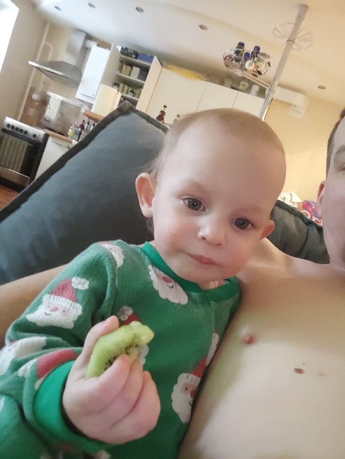 A young child, wearing green pajamas with Santa Claus patterns, is seated beside an adult. The child is holding a peeled kiwi in their left hand. The adult's chest and right arm are visible; they have light skin and two moles are visible on their chest. The background features a kitchen with appliances and white cabinets. The scene appears to be a casual snapshot taken indoors.