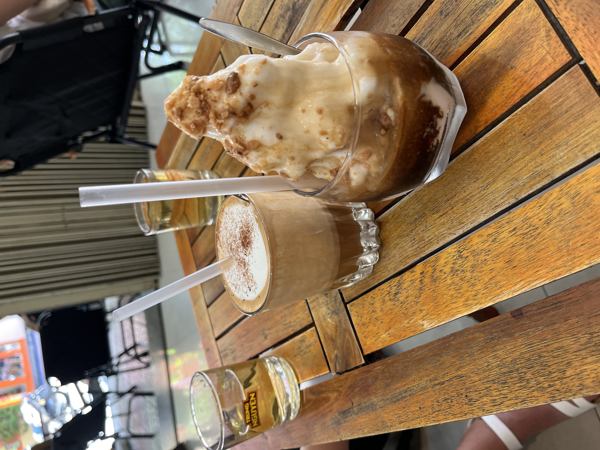 A top-down, tilted shot reveals a wooden slat table in an outdoor setting, likely in Đà Nẵng, Vietnam. On the table are three glass containers. At the top is a large, clear glass filled with a brown liquid, topped with a substantial amount of white foam and sprinkled with brown pieces, accompanied by a silver spoon. Below, a shorter, clear glass holds a layered coffee drink with a white frothy top dusted with brown powder, with a straw inserted. Beside these two drinks, a small glass with a gold-colored liquid, potentially tea, is present with a straw. A chair and elements of a street scene are visible in the background.