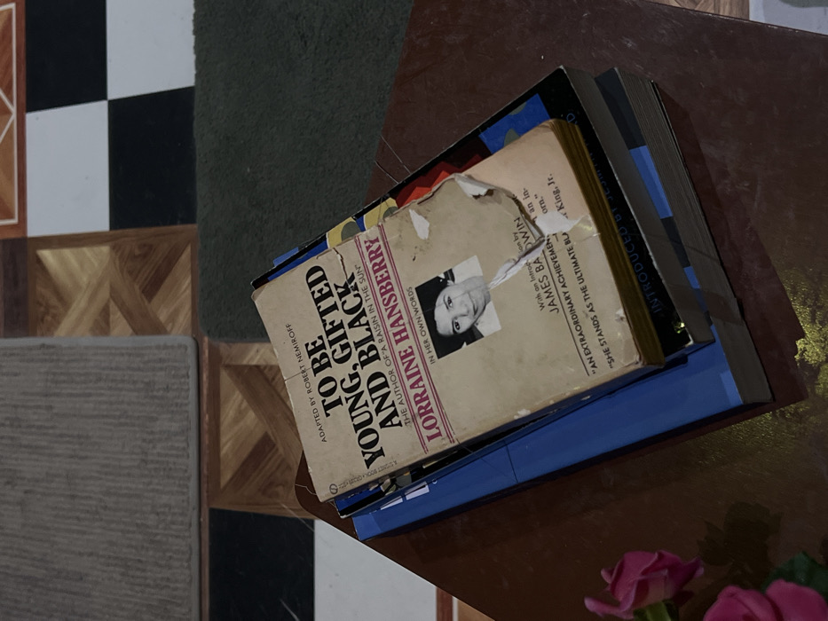Located in Paris, France, the image depicts a stack of books resting on a polished, dark brown surface, likely a table. Dominating the stack is a visibly aged and worn paperback copy of "TO BE YOUNG, GIFTED AND BLACK" by Lorraine Hansberry, adapted by Robert Nemiroff. The book's cover features a black and white portrait of Lorraine Hansberry; the lower right quadrant of the cover, beneath her portrait, exhibits significant tears and damage, revealing the pages beneath. Text visible on the cover includes "THE AUTHOR OF 'A RAISIN IN THE SUN'" and an introduction credited to "JAMES BALDWIN," along with a partially visible quote: "AN EXTRAORDINARY ACHIEVEMENT." Beneath this top book, at least two other books are stacked; the topmost of these has a prominent blue cover. To the immediate left of the book stack, a small cluster of artificial pink flowers, possibly roses, rests on the same dark brown surface. The background reveals an indoor floor composed of varied patterns: light brown geometric wood-like sections, a checkerboard pattern of black and white squares, and two floor mats—one light gray and textured in the upper left, and another dark green and plush in the upper right. The scene is illuminated by ambient, indirect lighting.