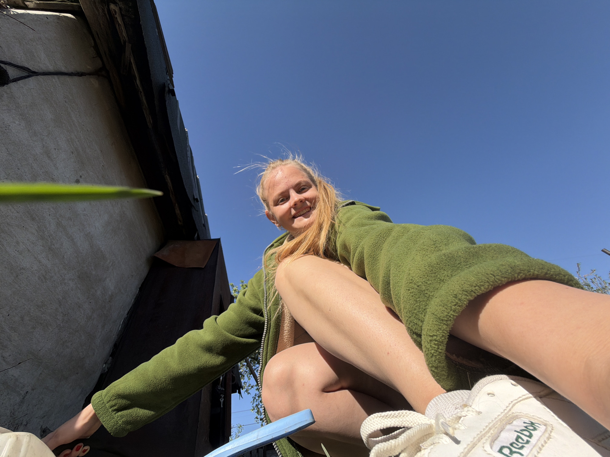 A low-angle shot shows a Caucasian woman with blonde hair, wearing a green fleece jacket and white Reebok sneakers. She is squatting outdoors with a clear blue sky visible above. The left side of the frame features a building's exterior wall. A blade of grass extends into the frame from the left. The woman is smiling at the camera. The location is Chaikovsky, Russia.