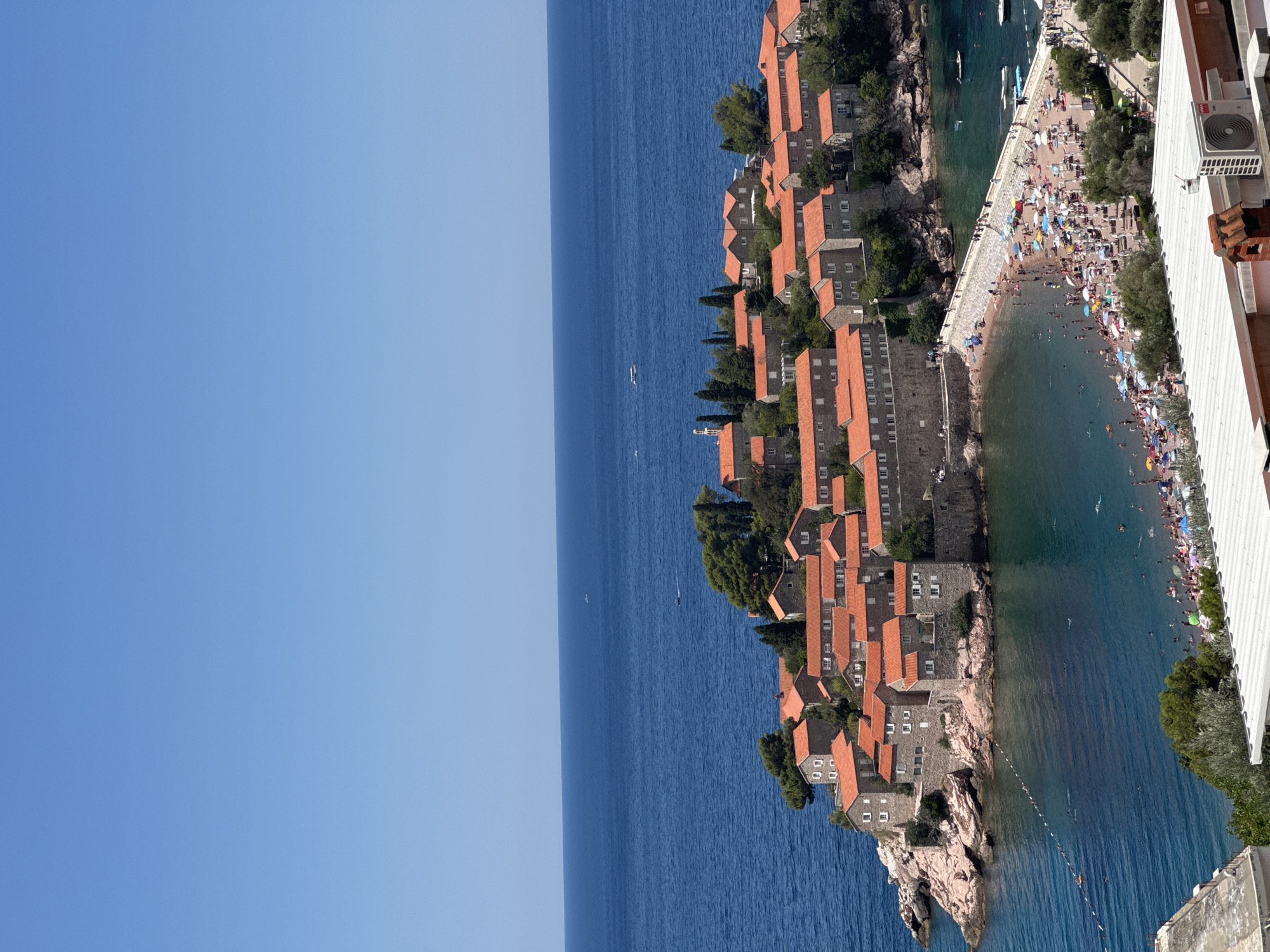This image captures the picturesque island of Sveti Stefan, Montenegro, a popular tourist destination. The island, connected to the mainland by a narrow causeway, is adorned with a cluster of stone buildings topped with red tile roofs, nestled amidst lush green trees. The causeway itself is bustling with activity, as numerous people are seen walking along it, enjoying the beach with colorful umbrellas and loungers spread across the sand. The clear blue waters of the Adriatic Sea surround the island and the beach, with several small boats and swimmers dotting the seascape. The sky above is a vibrant, cloudless blue, indicating a sunny day. The overall scene portrays a serene and idyllic coastal resort.