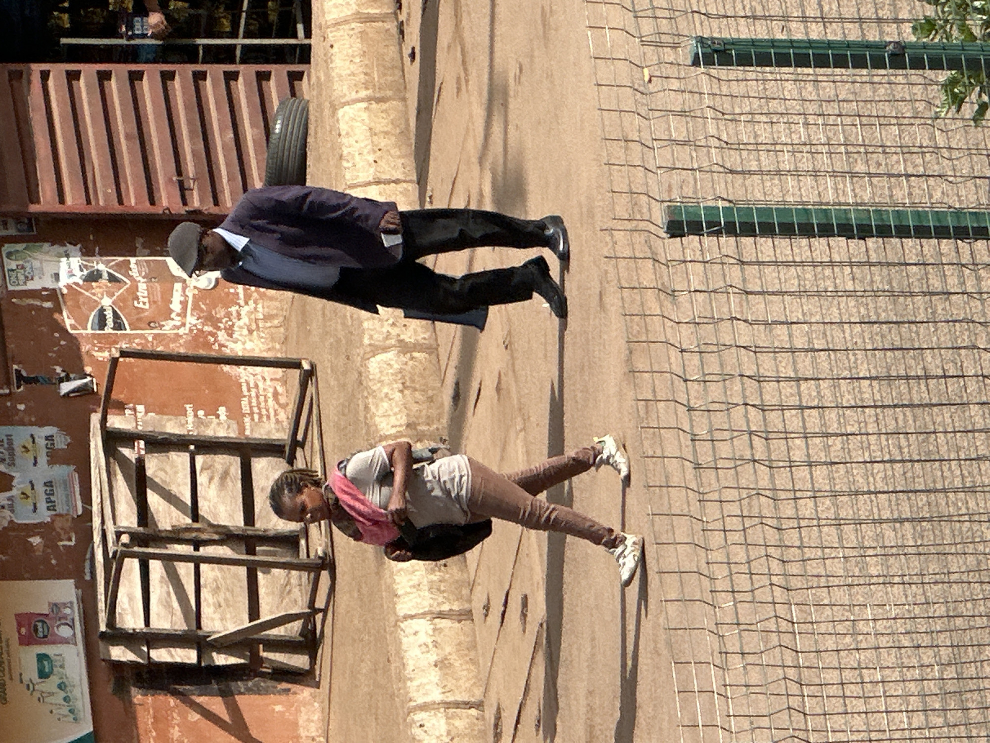 The image shows a street scene in Bukuru, Nigeria. In the foreground, a chain-link fence runs horizontally across the frame. Behind the fence, two people are walking on a paved surface. On the left, a young woman with braided hair walks towards the right, carrying a dark bag over her left shoulder and wearing a pink scarf around her neck, a grey t-shirt, and brown pants. She is wearing white and grey athletic shoes. On the right, an older man in a dark suit jacket, light blue shirt, and dark pants walks towards the right, wearing a dark flat cap and dark dress shoes. He is carrying a dark bag in his left hand. In the background, there are buildings with weathered walls, some with signs and posters. A large wooden structure, possibly a market stall or a shelter, stands to the left of the man. A tire is visible near the right side of the frame, leaning against a building with red horizontal louvers. The ground is paved and appears to be dusty. The lighting suggests it is daytime, with shadows cast by the people and structures.