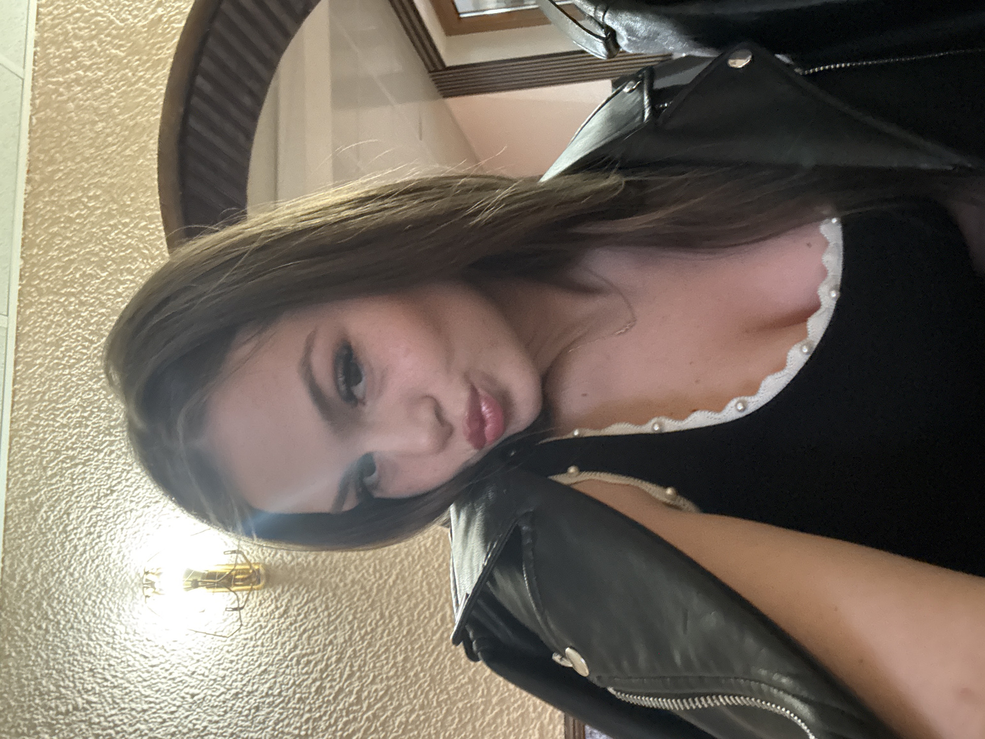A selfie of a woman with brown hair wearing a black top with a white pearl-style trim and a black leather jacket. The background includes a textured wall and a portion of a framed mirror with dark wood trim. There is no information present indicating a location in Parvomay, Bulgaria.