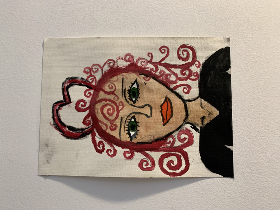 A vertically oriented, rectangular canvas painting is affixed to a plain, light-colored wall. The painting depicts a stylized, frontal portrait of a female-presenting figure from the chest up. The figure has light beige skin, large, wide-set green eyes with black pupils and outlines, thin black arched eyebrows, a simplified black line nose, and bright orange lips outlined in black. Small black marks are present on the right cheek. The figure's hair is a mass of bright red spirals and curls, with a prominent red heart shape outlined in black sitting atop the head. The figure wears a black V-neck top or dress. The canvas surface shows visible texture. The top left corner of the canvas exhibits minor dark smudges, and the wall behind it also has faint dark marks. A slight shadow is cast along the left edge of the painting. The scene is located in Vozdovac Urban Municipality, Serbia.