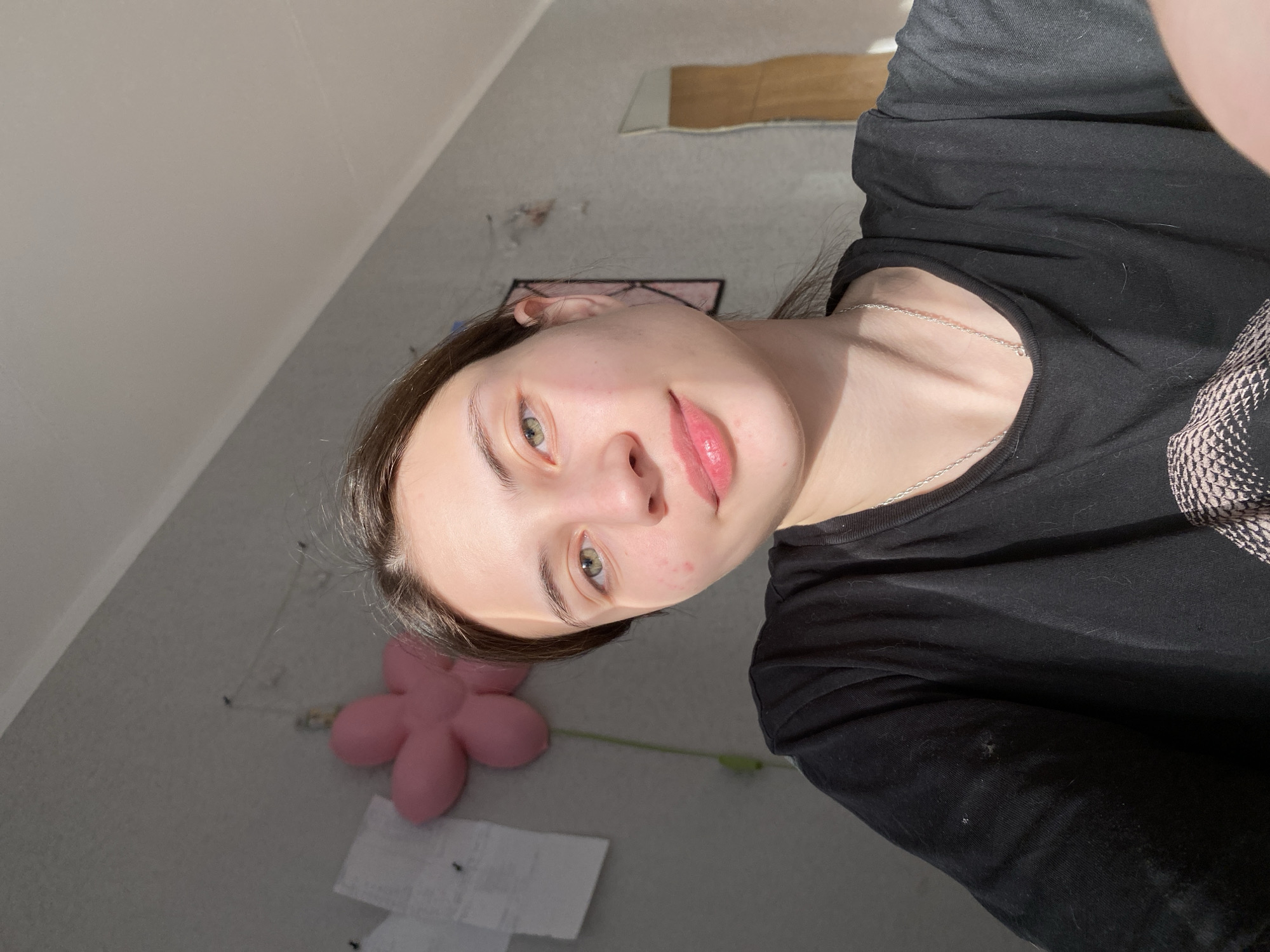 A pale-skinned young woman with dark hair and green eyes is centered in the frame, angled from the lower right to the upper left. She is wearing a black t-shirt and a silver necklace. The background includes a light gray wall with a pink flower-shaped object and several white papers affixed to it. The image appears to be taken indoors, possibly in a bedroom. The location in Votkinsk, Russia is not discernible from the image.