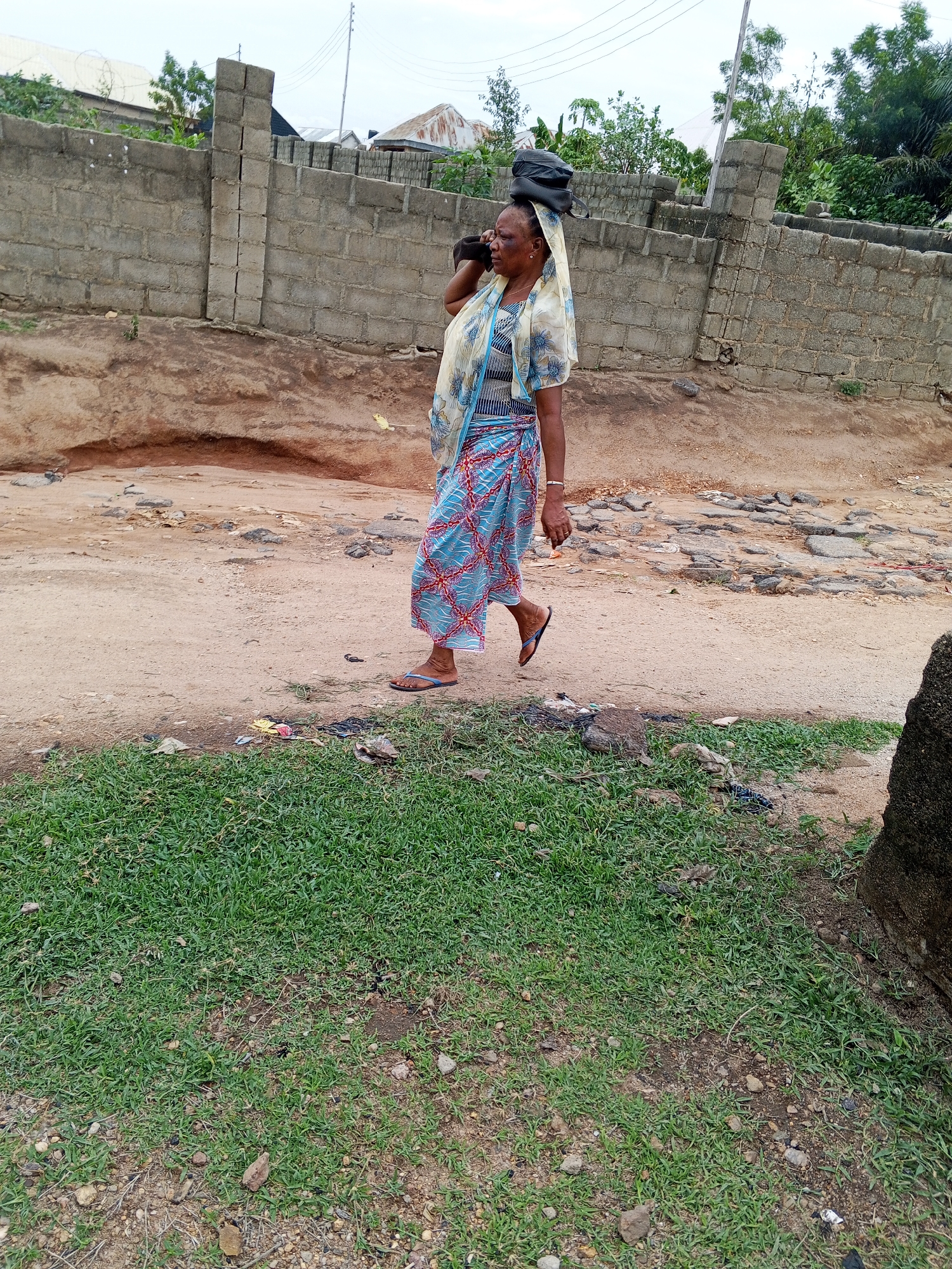 An adult woman is walking on a dirt road in Zango-ndaji, Nigeria. She carries a dark-colored load on her head, held in place by a patterned headscarf. She is wearing a blue, red, and white patterned wrap skirt and a light-colored patterned top with a blue striped shirt underneath. She also has on a bracelet. The road is uneven with patches of exposed rock and dirt, and there is trash scattered at the edge of the road near a patch of green grass. In the background, there is a grey cinder block wall, some vegetation, and a few buildings. Utility poles are visible in the distance.