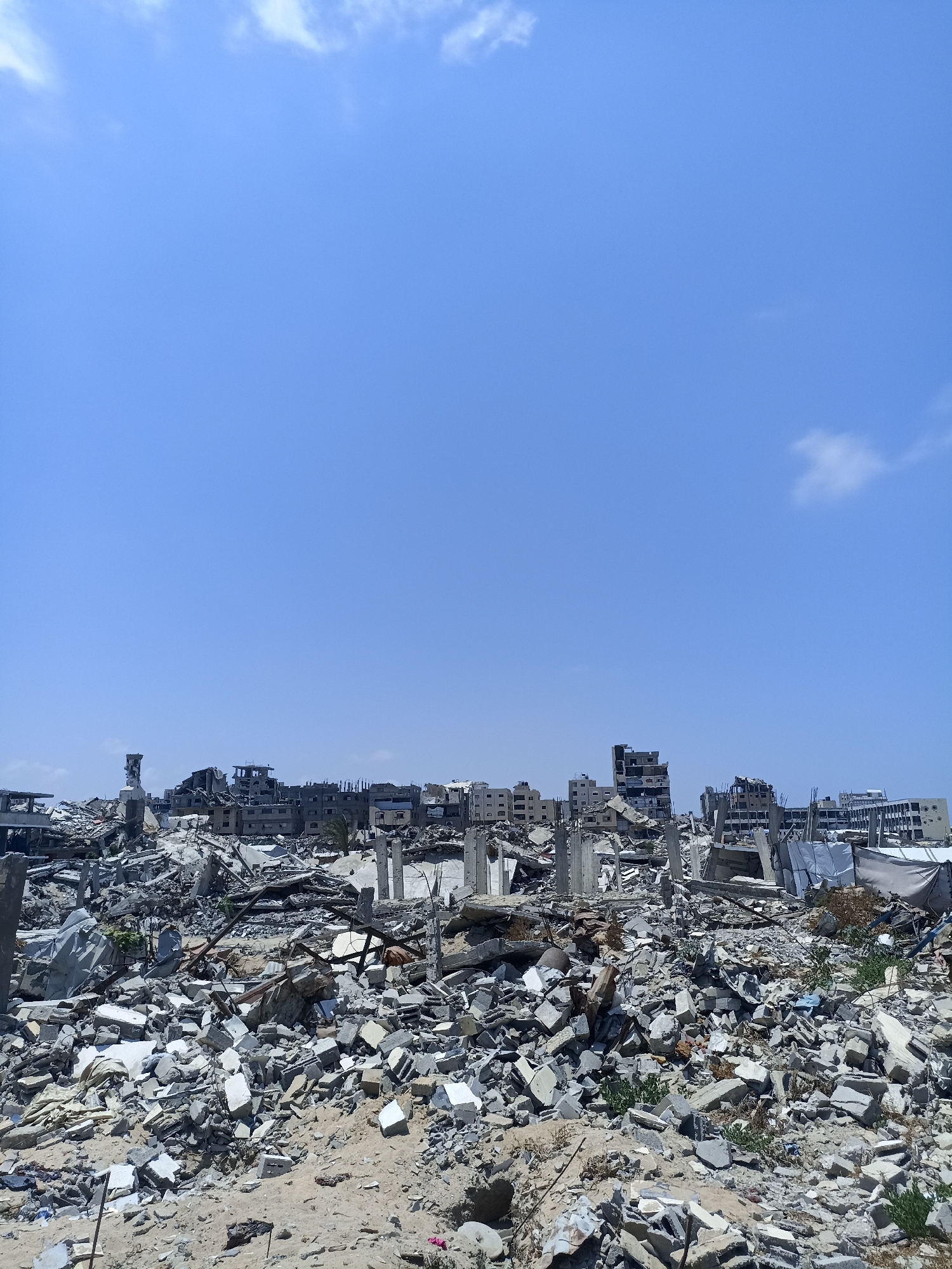 The image depicts a scene of widespread destruction in Jabalia, Palestinian Territories, under a clear blue sky with scattered white clouds. The foreground is dominated by a vast expanse of rubble, composed of shattered concrete, bricks, rebar, and other building materials. Among the debris, remnants of building structures are visible, including concrete pillars and fragmented walls. In the background, a cityscape of partially destroyed multi-story buildings can be seen, some with gaping holes and missing sections. The overall impression is one of devastation and the aftermath of conflict. There are no people or animals visible in the image. The environment is characterized by the ruins of what were once urban structures.