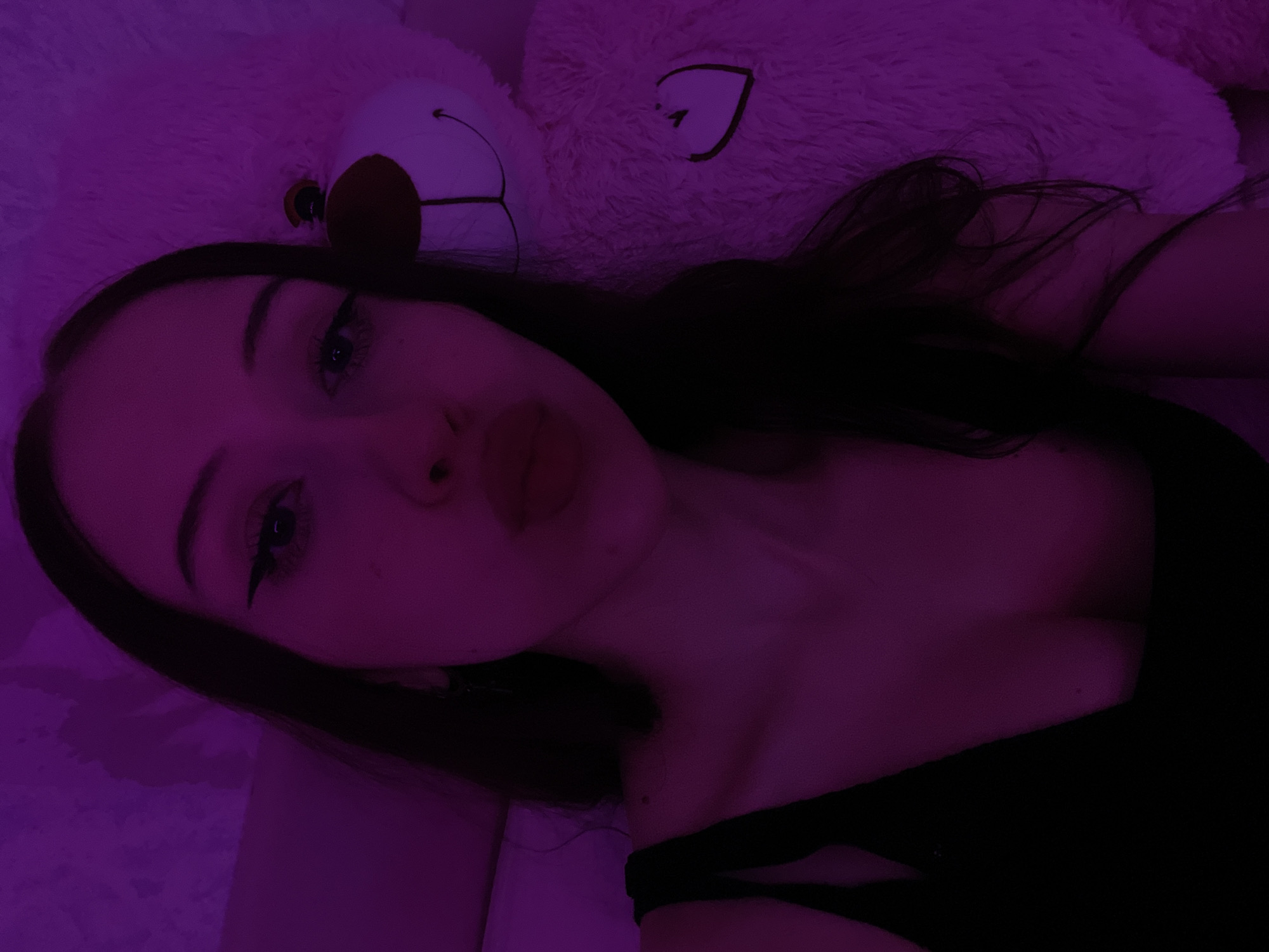 A young woman with dark hair is taking a selfie in a dimly lit room, illuminated by purple light. She is wearing a black top. Next to her, on the right, is a large, fluffy white teddy bear. The bear has a stitched smile and a heart design on its chest. The background is a textured wall or surface. There is no information to suggest this photo was taken in Votkinsk, Russia.