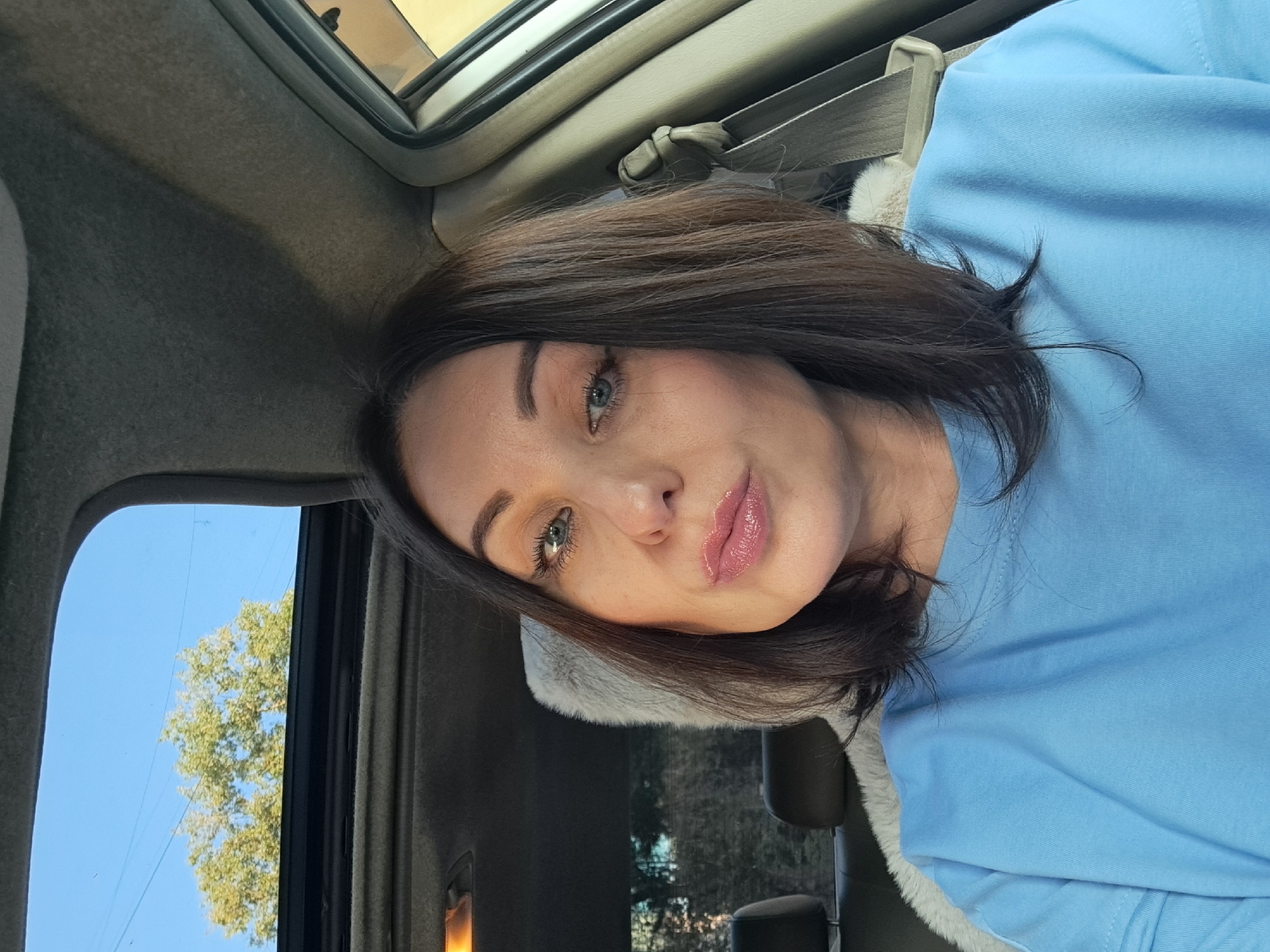 A selfie taken from inside a car shows a woman with brown hair and blue eyes looking at the camera. She is wearing a light blue t-shirt and has her lips slightly pursed. The car's sunroof is open, revealing a clear blue sky with a tree in the background. The interior of the car is visible, including the dashboard, a seatbelt, and a fluffy seat cover. The lighting suggests it is daytime. The location is not specified in the image.