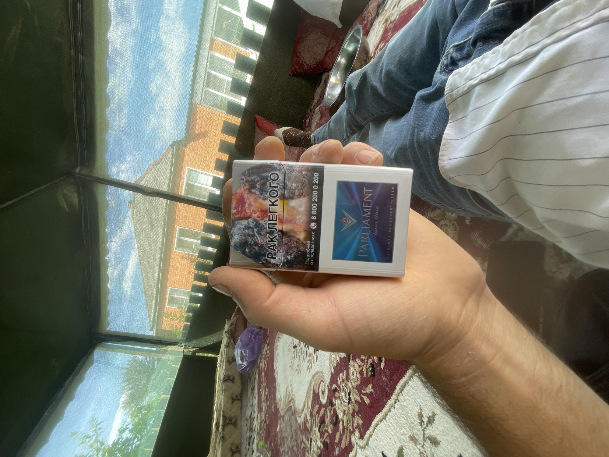 A person's left hand holds a pack of Parliament Aqua Blue cigarettes. The cigarette pack is white with blue and gold accents, featuring a graphic of lungs on the front with Russian text. The hand is positioned in the foreground, with the pack angled towards the camera. In the background, a person's legs are visible, clad in blue jeans, with a metallic bowl resting on their lap. The scene appears to be set outdoors, possibly on a patio or porch, with a patterned rug visible on the ground. Beyond the immediate area, a brick house with windows and a roof is discernible, with a blue sky with scattered clouds overhead. The overall environment suggests a relaxed, casual setting.