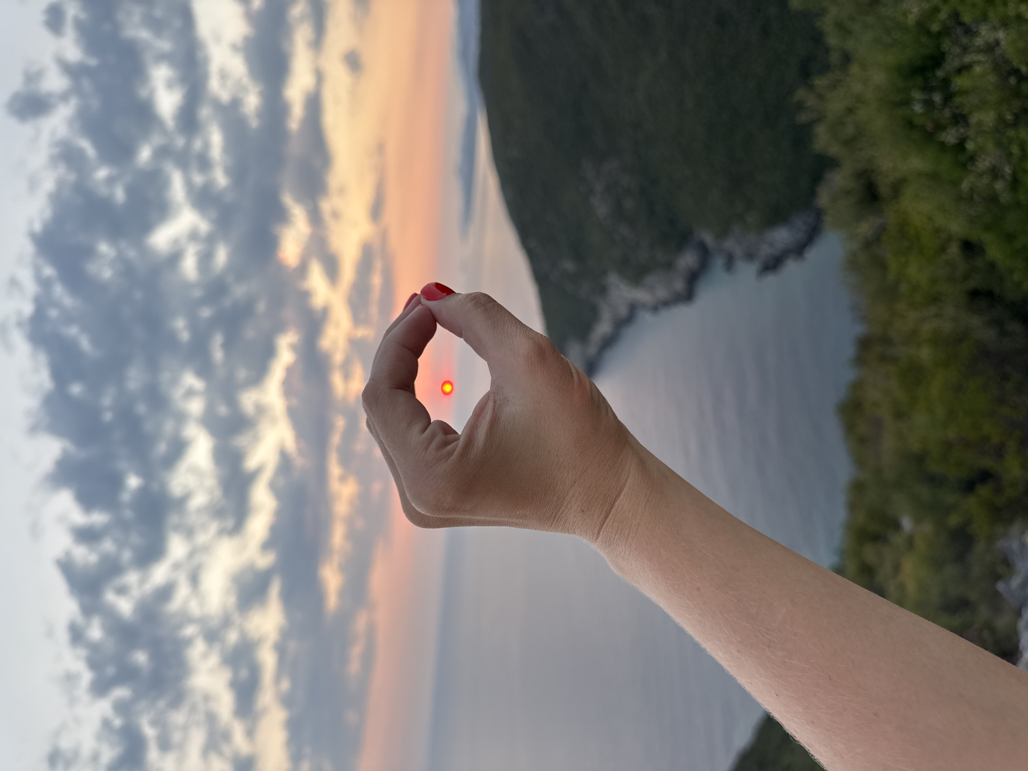 A person's hand with red nail polish is held up in the foreground, forming an "O" shape with their thumb and index finger. The setting sun, a bright orange orb, is positioned within this "O" shape. The background depicts a serene coastal landscape with the sea stretching to the horizon, a hazy sunset with orange and pink hues, and a green, hilly coastline with trees. The image appears to be taken from an elevated viewpoint overlooking a bay. The specific location within the city of Zagora, Montenegro, is not discernible from the image itself.