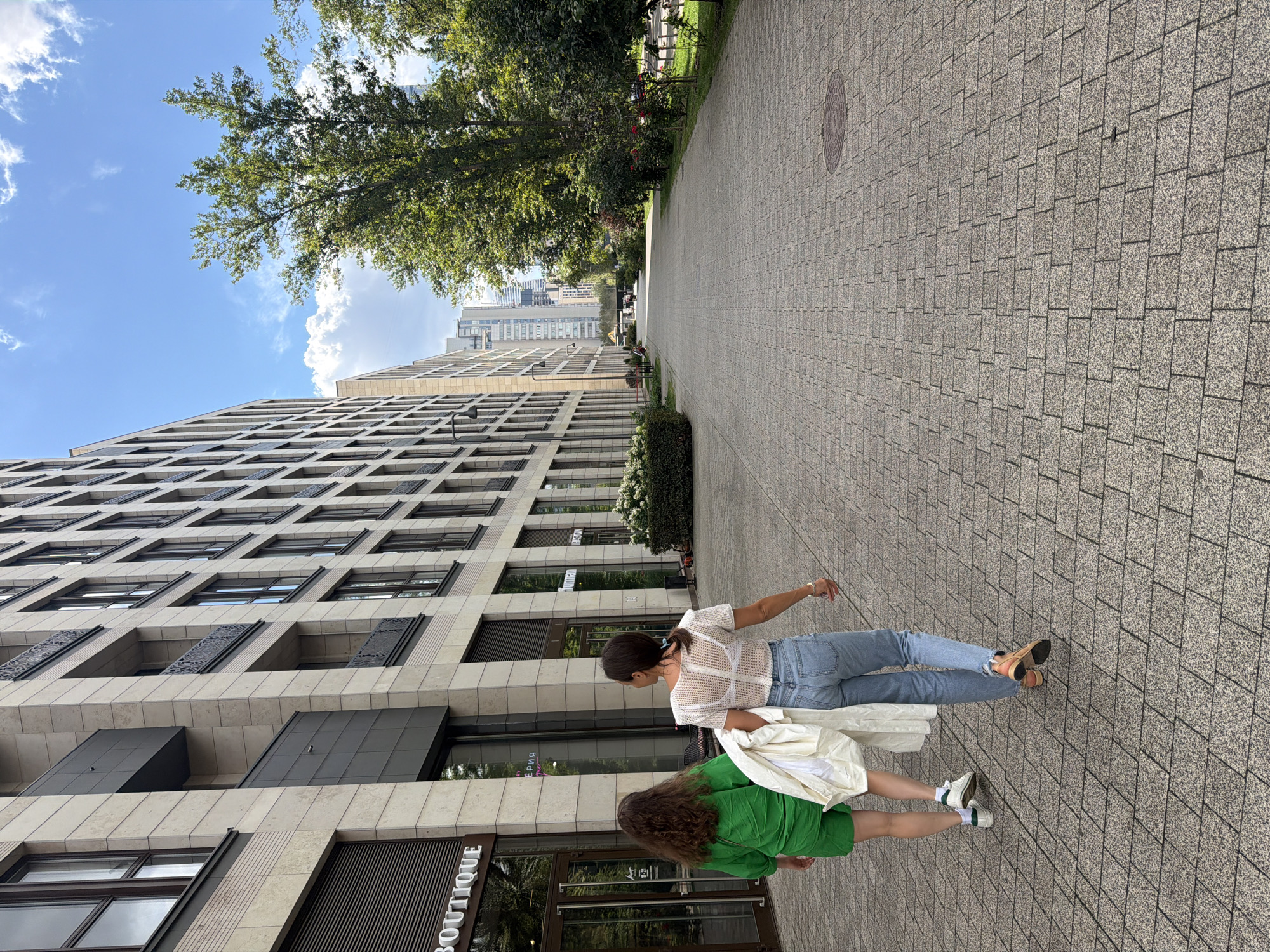 Two women walk away from the camera on a paved walkway alongside a modern building in Moscow, Russia. The woman on the left wears a green outfit and has curly brown hair. The woman on the right wears a white sheer top, blue jeans, and carries a white garment over her arm. The building to their left features a light-colored stone facade with numerous windows and balconies. On the right side of the walkway, lush green trees and bushes line the path, with a distant view of skyscrapers. The sky is blue with scattered white clouds. The paving stones are rectangular and arranged in a geometric pattern.