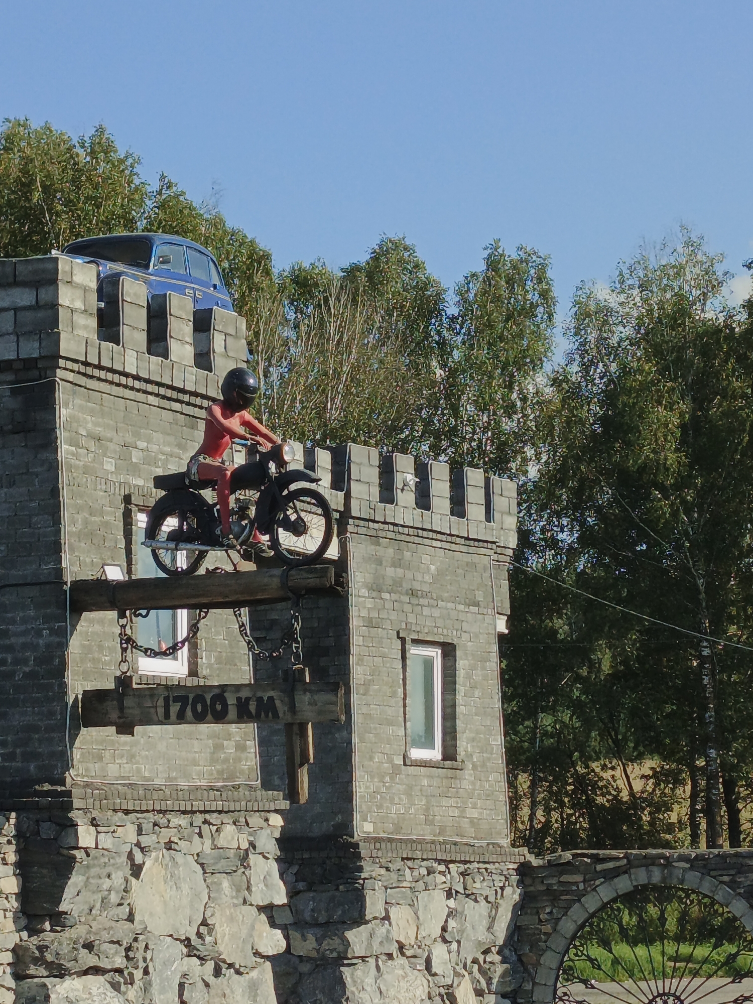 In Berezovyi Most, Russia, a unique display features a motorcycle with a rider perched atop a brick structure resembling a castle. The motorcycle, a vintage model, is positioned on a wooden beam extending from the structure. The rider, a mannequin, is wearing a red outfit and a black helmet. Below the beam, a wooden sign reads "1700 KM" and is suspended by chains. The structure itself is made of brick and stone, with a blue car placed on top of the castle-like crenellations. Trees and a clear blue sky form the background. The scene appears to be an outdoor attraction, possibly a roadside exhibit.