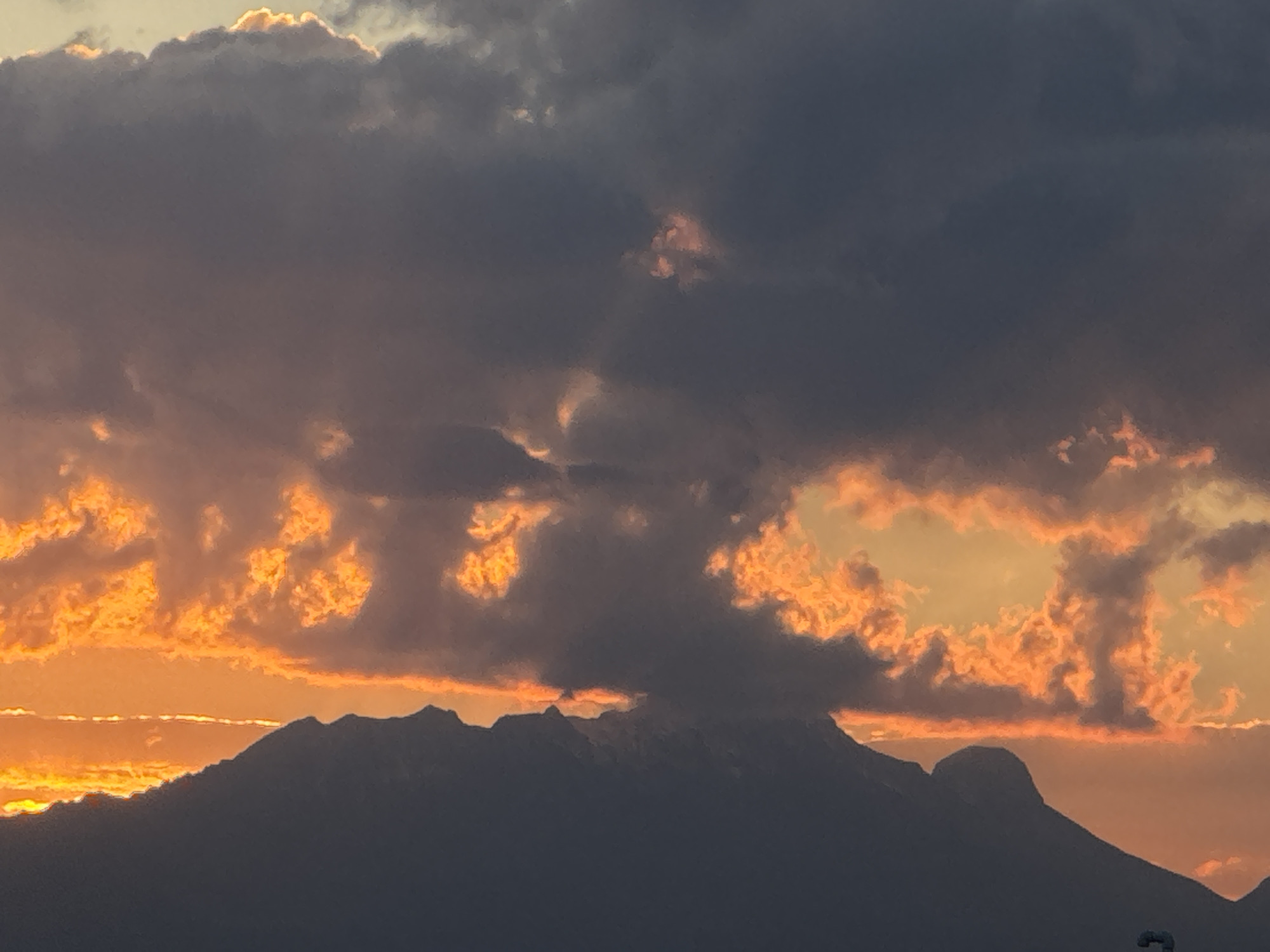 The image captures a dramatic sunset over a mountain range in Ocotlán, Mexico. Dark, silhouetted mountains dominate the lower portion of the frame. The sky is filled with dark, heavy clouds, some of which are lit from behind by the setting sun, creating patches of orange and yellow light. The overall scene is moody and striking, with a contrast between the darkness of the mountains and clouds and the bright colors of the sunset. No people are visible in the image.