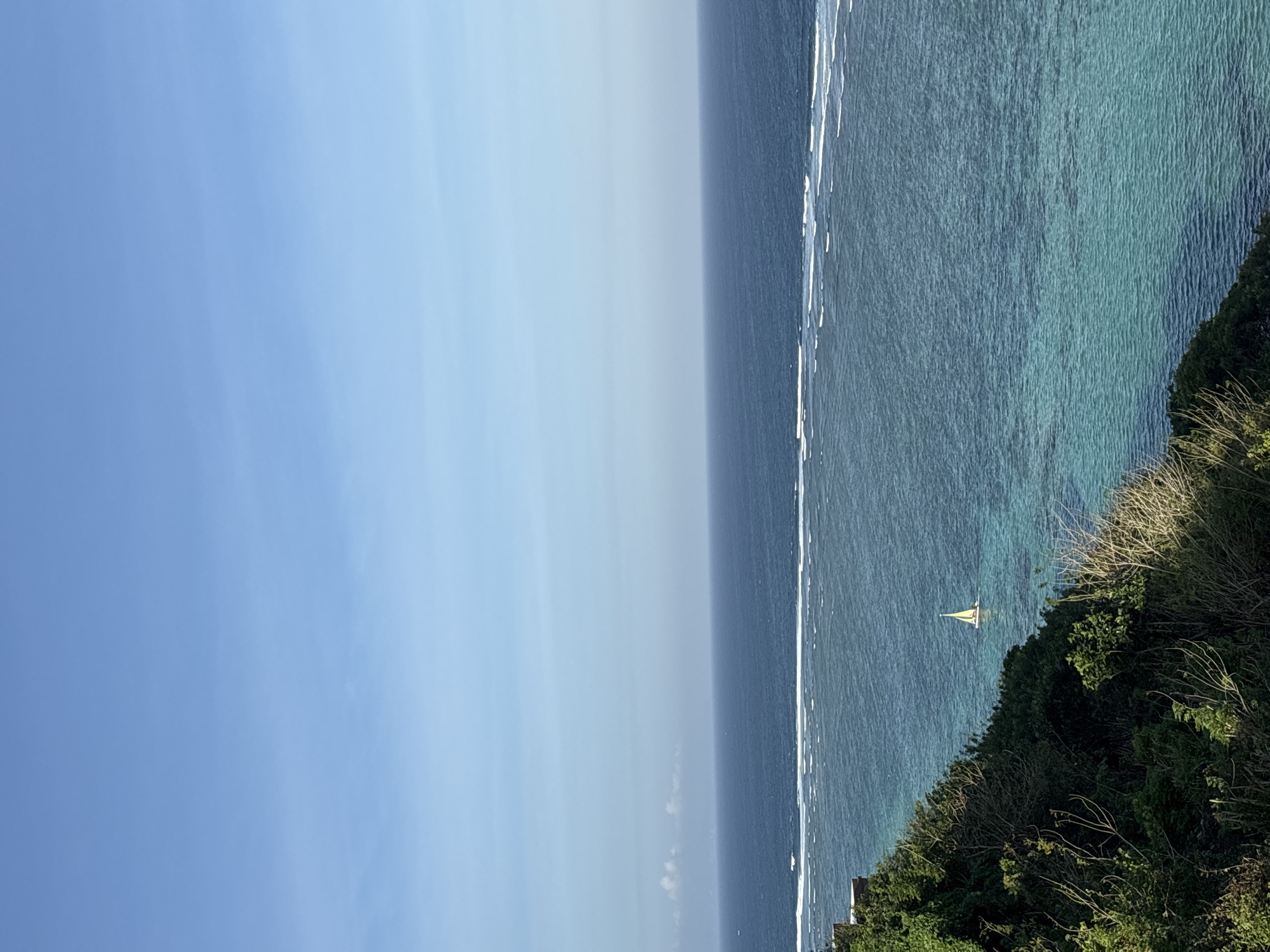 The image depicts a serene coastal scene in Kuta Selatan, Indonesia. The vast expanse of the ocean dominates the frame, with its surface shimmering under the daylight. A narrow strip of white surf runs parallel to the shore, marking the edge of a coral reef that lies just offshore. Beyond the reef, the water transitions into a vibrant turquoise, revealing the shallow depths and the underwater topography.
On the right side of the image, lush green vegetation forms a dense cliffside, with dry grasses and branches interspersed amongst the foliage. A small sailboat with a yellow sail is visible in the distance, gliding across the tranquil blue waters, adding a touch of life and movement to the otherwise still seascape. The sky above is a clear, soft blue, indicating fair weather and a pleasant time of day, likely late morning or early afternoon. The overall impression is one of peace and natural beauty, a typical view of the Indonesian coastline. There is no visible text in the image.