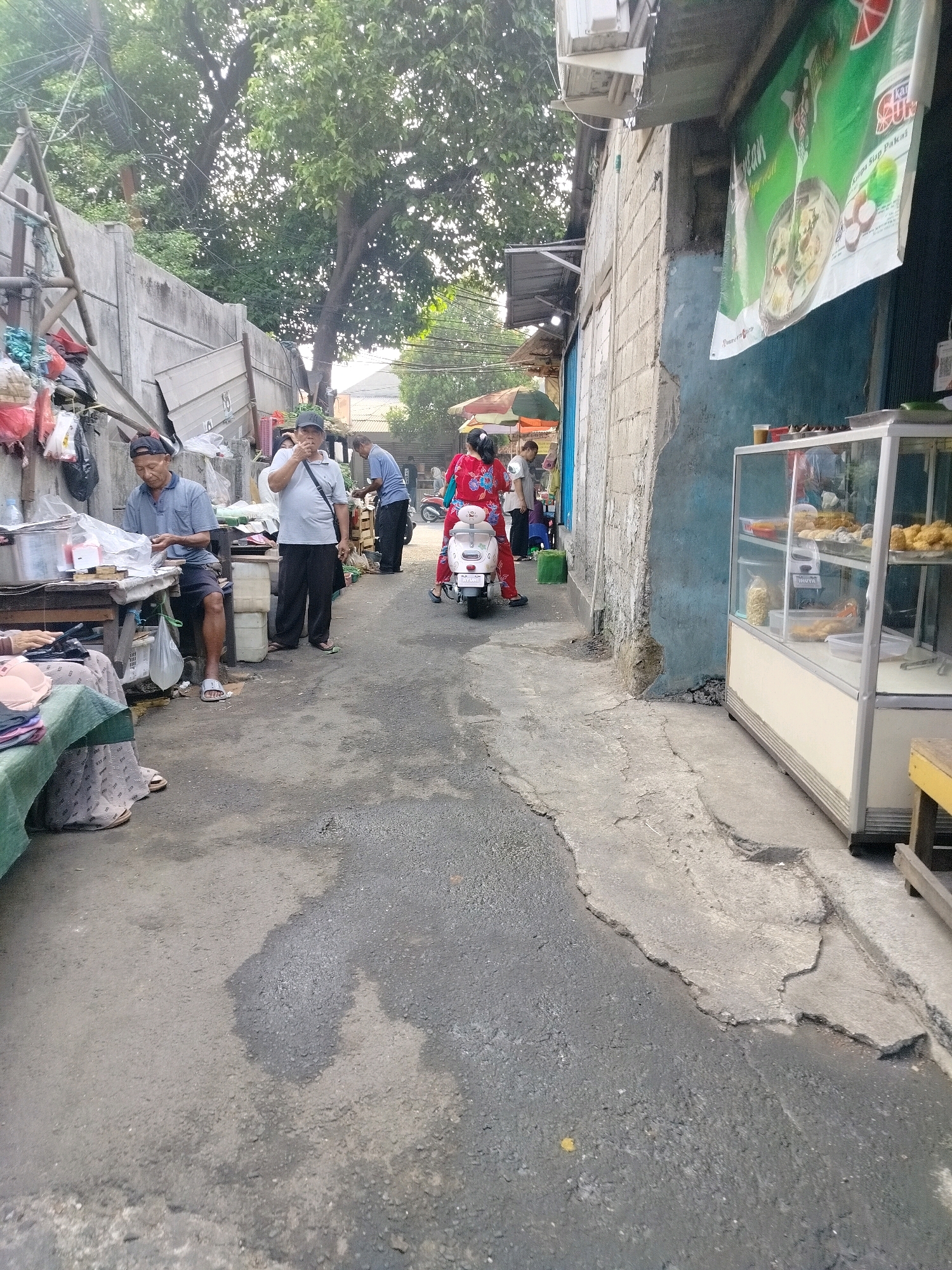 In a narrow, outdoor alleyway in East Jakarta, Indonesia, several individuals are engaged in various activities. The environment is characterized by weathered concrete walls, an asphalt ground with patches of damage and discoloration, and overgrown trees providing shade from above.
On the left side of the alley, a man wearing a blue polo shirt and dark shorts sits at a wooden table, appearing to be engaged in work or a transaction. Next to him, another man in a white polo shirt and dark pants stands, holding something to his mouth. Further down the alley, a man in a blue shirt and dark pants is seen from the back, seemingly interacting with a display.
In the center, a woman on a white scooter, wearing a red and white patterned outfit, is in motion, heading away from the viewer. Another woman in a similar red outfit stands near the scooter, also facing away.
Along the right side of the alley, a glass display case filled with food items stands against a building with a blue facade. A large green banner with text and an image of food hangs above the display. Overhead, electrical wires crisscross the sky, and parts of buildings and roofs are visible. The overall scene suggests a bustling local market or street vendor area.