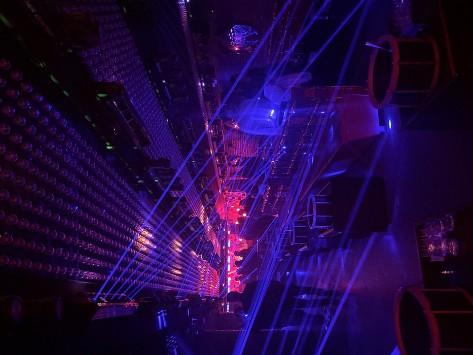 This image depicts the interior of a multi-level nightclub, specifically identified as Teatro Kapital in Madrid, Spain. The scene is dominated by dark ambient lighting, intensely illuminated by numerous blue laser beams crisscrossing the entire space, originating from unseen projectors. Ambient purple light also permeates the venue, while a vibrant red-lit stage or large screen is visible at the far end.
The ceiling is covered by a dense grid of small, reflective spherical elements, creating a visually textured surface. Large sound and lighting fixtures, some exhibiting small green indicator lights, are mounted overhead. The floor is dark and somewhat reflective.
In the foreground, two dark, cylindrical containers resembling drums are positioned; one holds a spoon and is placed near several clear glasses. A row of dark, low platforms or tables extends from the mid-ground towards the background, some also featuring smaller drum-like containers. Along the right side of the image, a long, reflective surface, likely a bar, is visible, with a textured wall, possibly brick, behind it that includes a circular, reflective accent.
Multiple indistinct human figures are present, appearing as dark silhouettes against the bright laser and stage lighting, suggesting a social gathering. One figure in the mid-ground on the right is partially illuminated by blue light while leaning against a surface. No specific actions or direct interactions are clearly discernible due to the low light and depth of field.