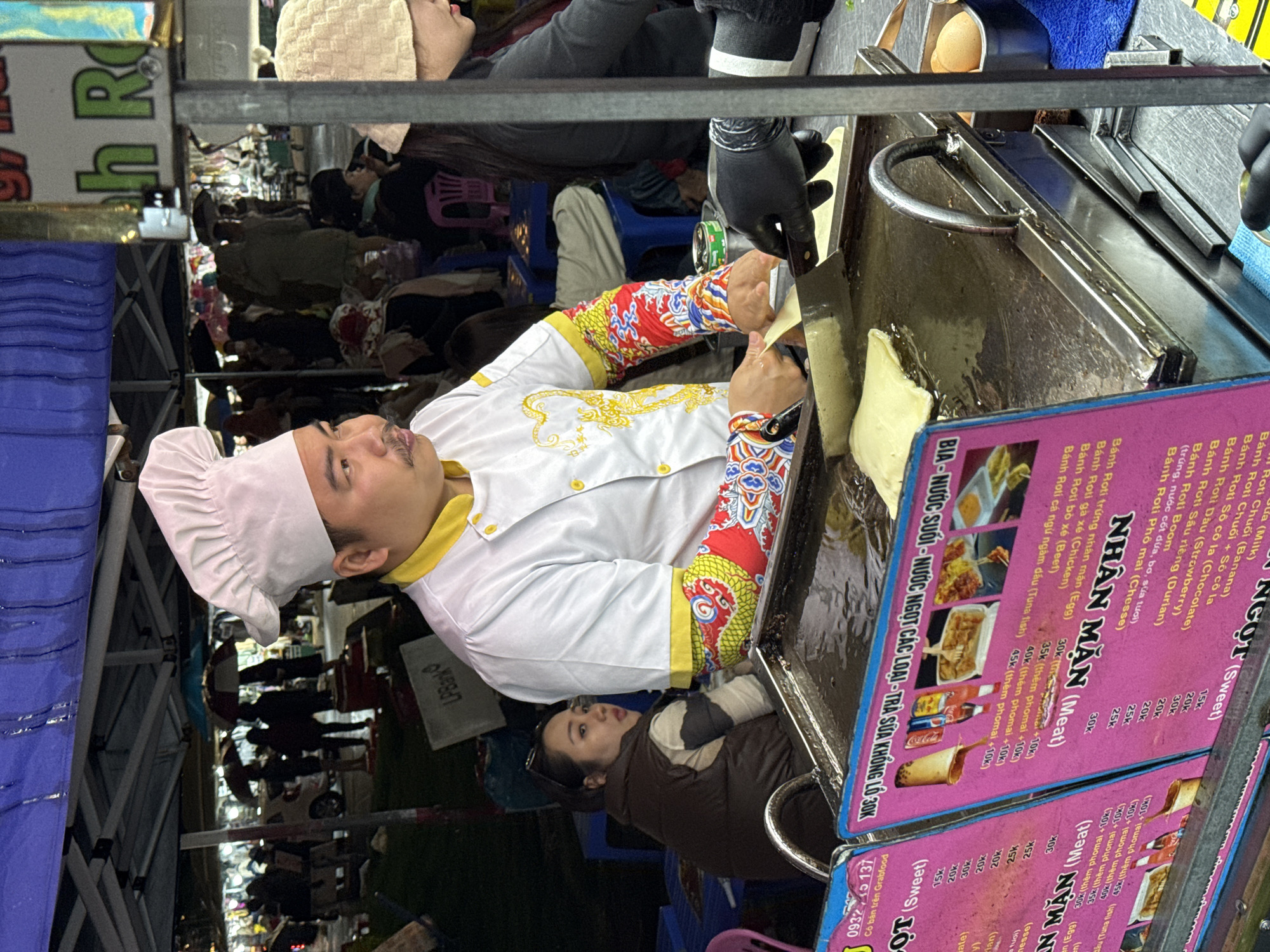 The image shows a street vendor in Da Lat, Vietnam, preparing and selling Banh Roti. A chef in a white uniform and hat is cooking on a hot plate. A price list in Vietnamese is visible, advertising various sweet and savory Banh Roti options. A woman is observing the chef. There are other people in the background, indicating a crowded area.