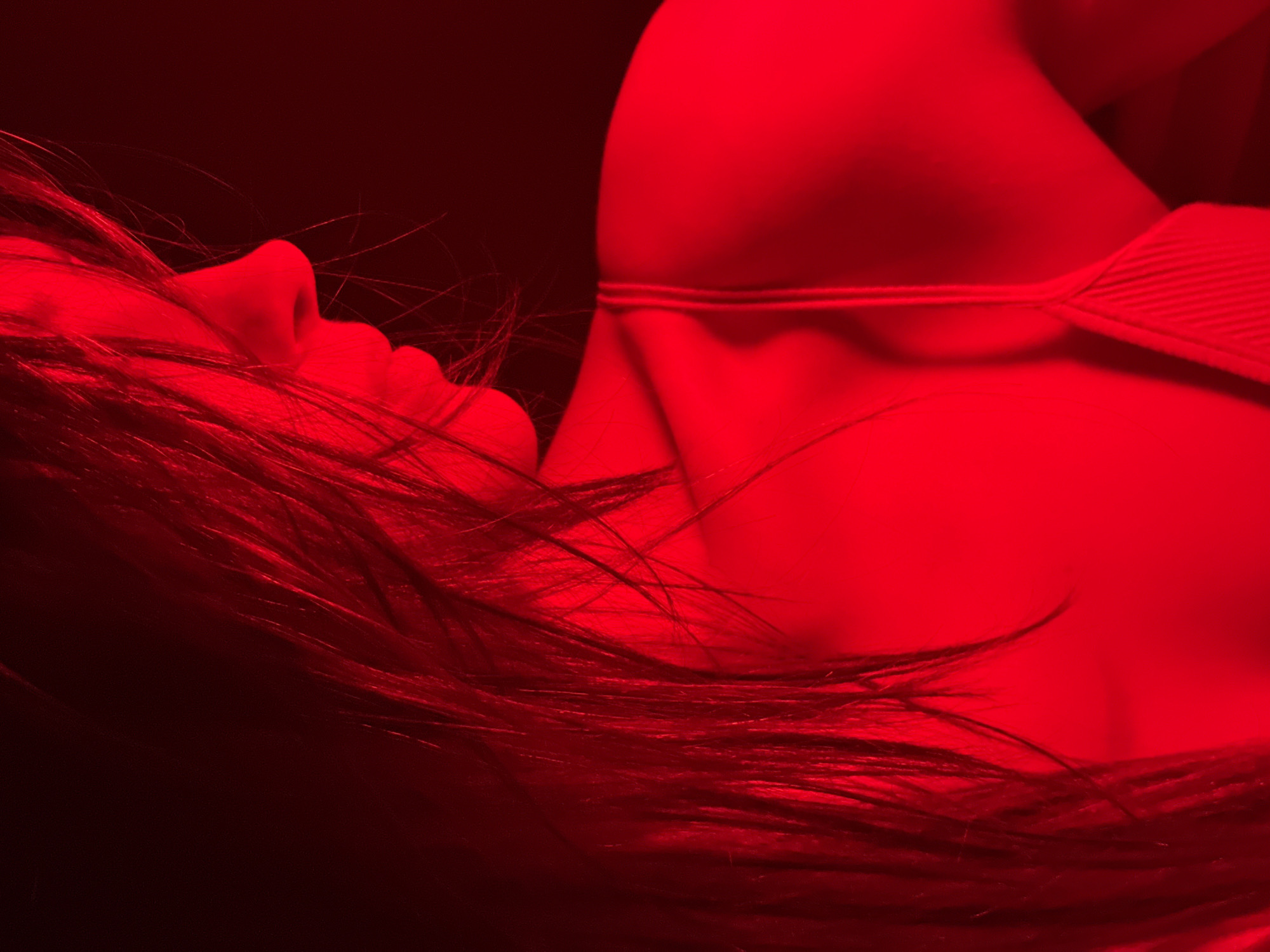 The image shows a person lying down, presumably female, illuminated by a red light. The person's face is visible in profile, with their hair splayed around their head. Part of their upper body is visible, including what appears to be a strap of clothing. The background is dark and indistinct.
The location in the city of Chaikovsky, Russia, is not discernable from the image.