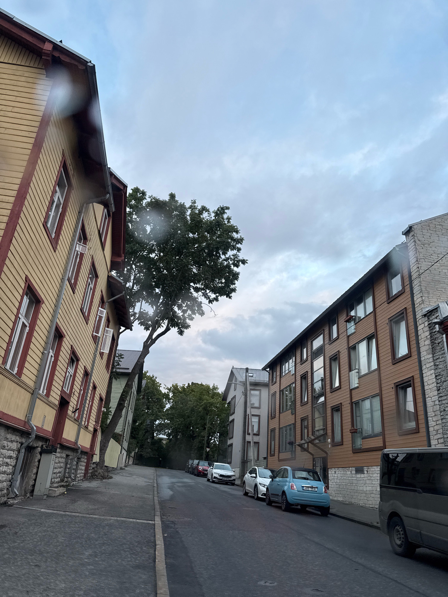 The image depicts a street scene in Tallinn, Estonia, characterized by a mix of architectural styles. On the left, a traditional wooden house with yellow siding and red trim leans precariously, its facade angled in perspective. The street itself is paved with cobblestones in the foreground and asphalt further down. Several cars are parked along the street, including a white sedan, a silver hatchback, and a small blue Fiat 500 with a license plate that reads "465 M-HK". In the background, a modern brown apartment building with numerous windows stands to the right, contrasted by a stone facade on its rightmost section. The sky is overcast with clouds, suggesting a potentially damp or cool day. A large tree with green foliage grows at an angle, appearing to sprout from behind the yellow wooden house. The overall atmosphere is quiet, with no visible people or active interactions.