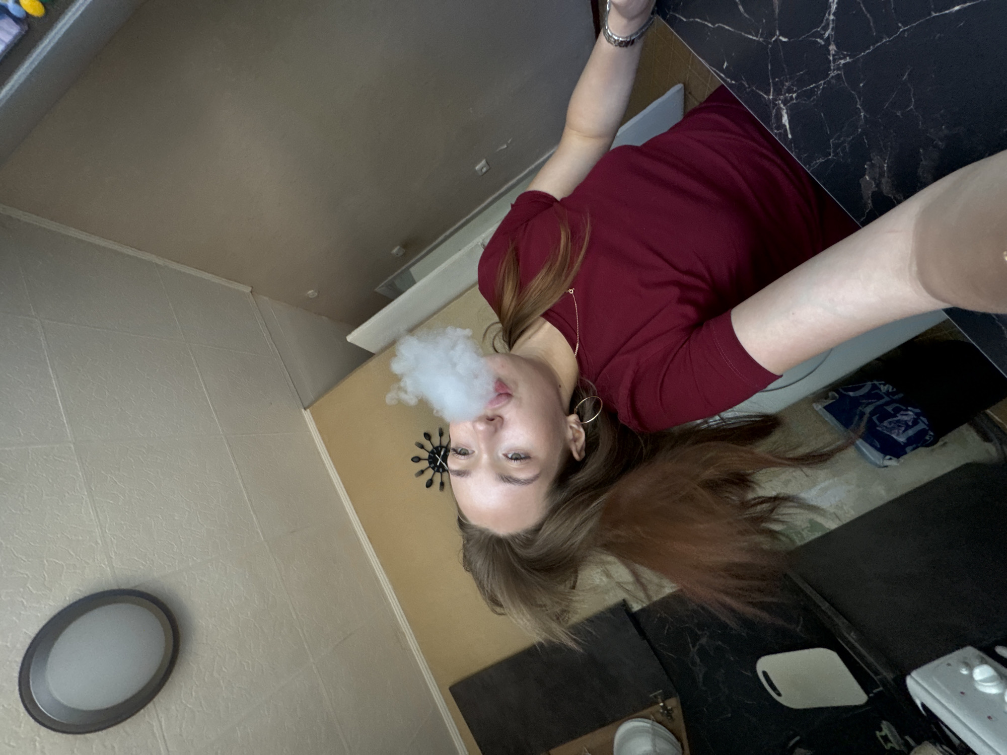 A young woman with long, auburn hair is seen exhaling a cloud of vapor. She is wearing a dark red t-shirt, a delicate necklace, and hoop earrings. Her head is tilted back, and her eyes are open. The background appears to be a kitchen, with a ceiling light fixture, a clock on the wall, and various kitchen items visible. A portion of a black countertop with a marble pattern is in the foreground. The scene is indoors, likely in a residential setting. The lighting suggests it is daytime, possibly from natural light entering the room.