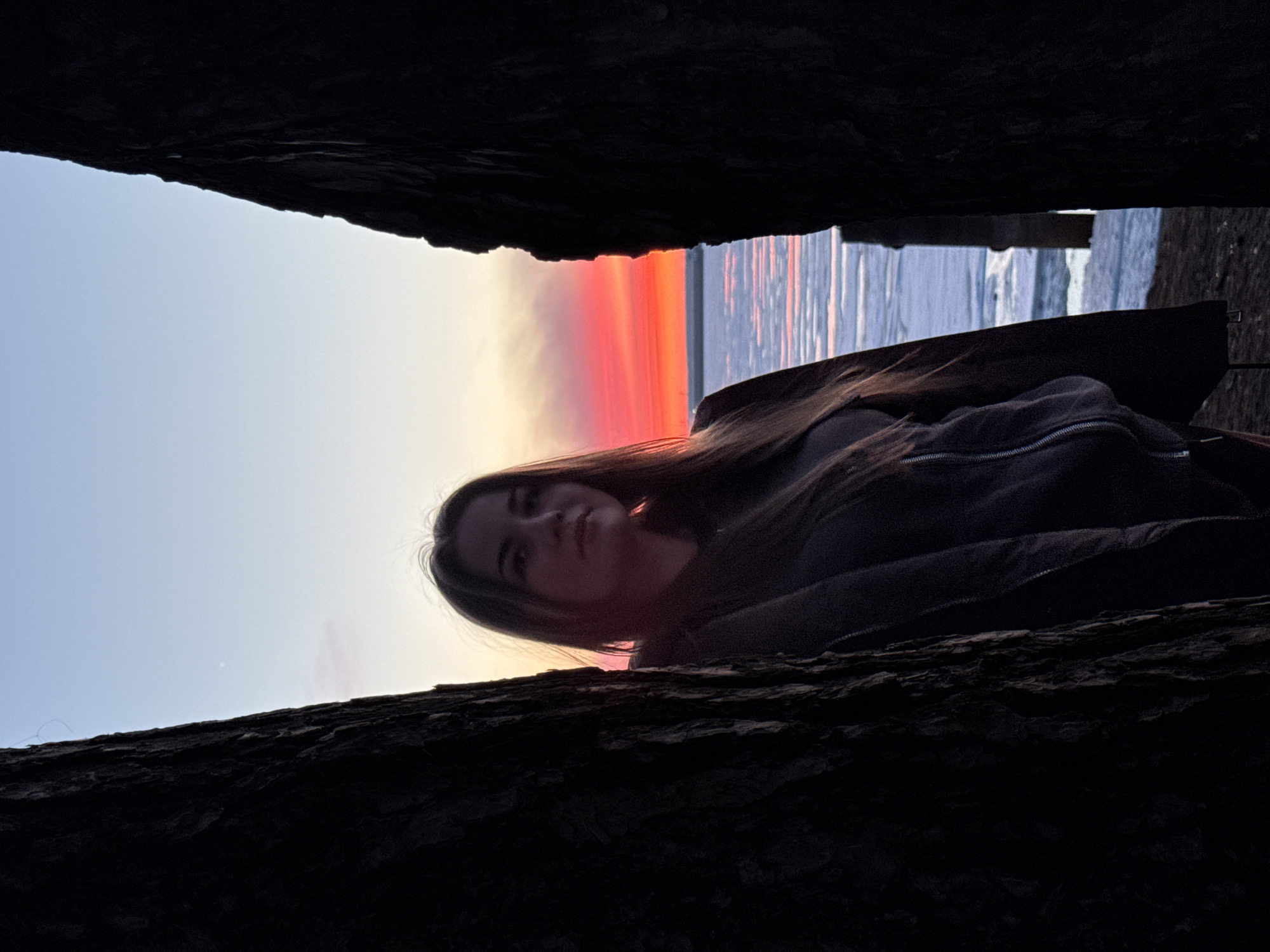 A person with long, dark hair lies on a dark surface, possibly a tree trunk, positioned between two dark masses. The background shows a clear sky transitioning to a red-orange sunset, which is reflected in water, likely a lake or river. The location is Chaikovsky, Russia.