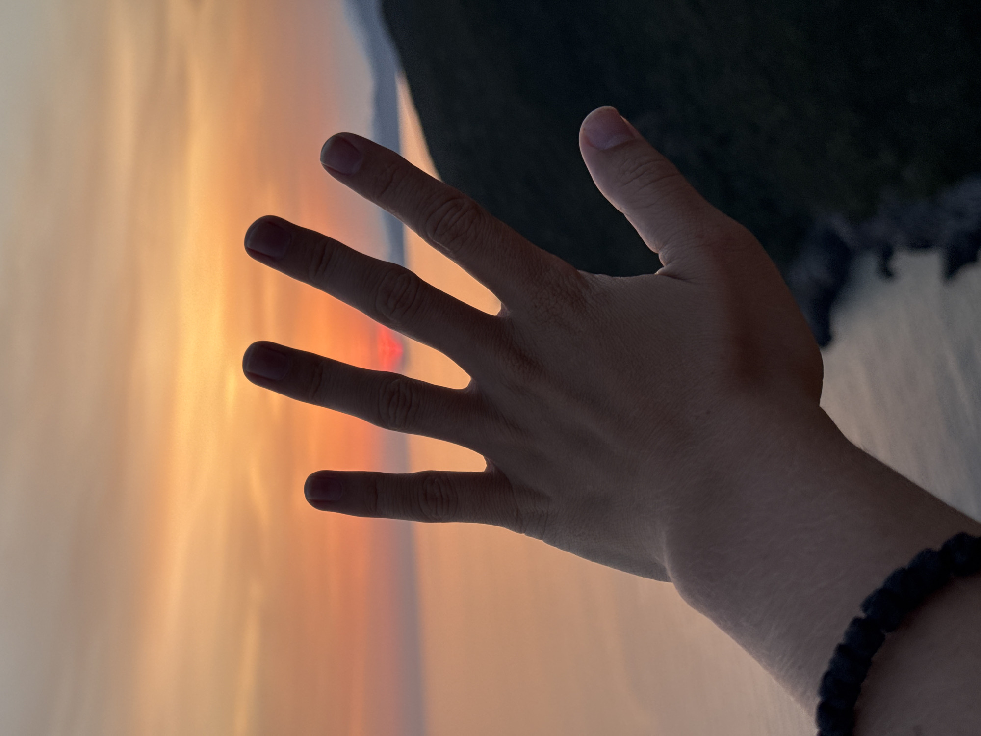 A close-up shot captures a person's right hand, palm facing forward, extended towards a sunset over the Adriatic Sea. The hand, adorned with a black beaded bracelet on the wrist, is silhouetted against the vibrant orange and yellow hues of the sky. The sun, a small, glowing disc, is partially obscured by the fingers. The background shows a dark, mountainous coastline on the right and the calm sea reflecting the sunset colors on the left. The scene is set in Zagora, Montenegro.