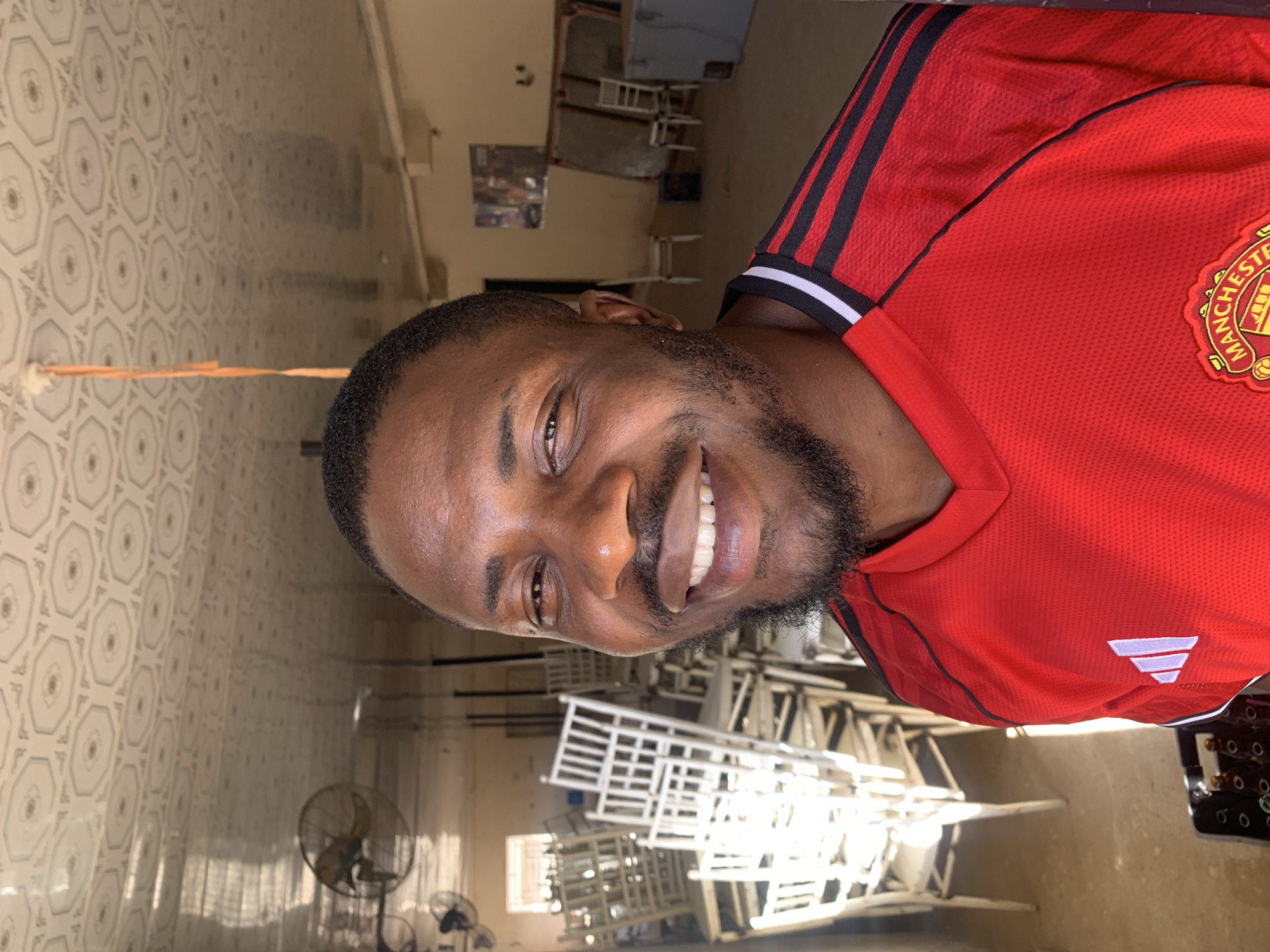 A medium shot depicts a smiling Black man with a beard and short hair wearing a red Manchester United soccer jersey with black stripes on the shoulders. The background features a room in Kabong, Nigeria, with white metal bed frames stacked against a wall. An orange cord is taped to the tiled wall. There are fans in the corner of the room.