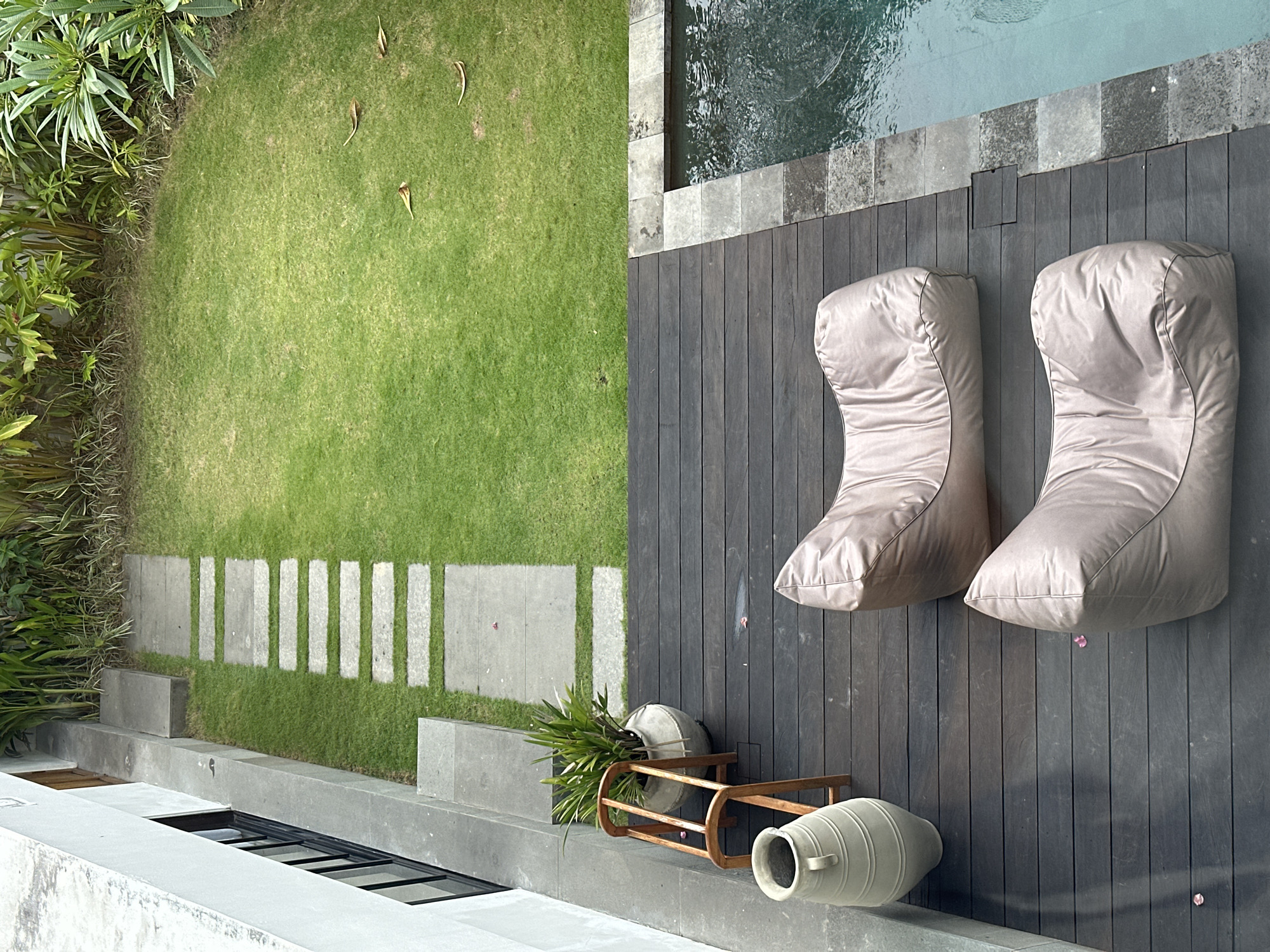 Captured from an elevated perspective, the photograph presents a portion of a luxurious outdoor area in North Kuta, Indonesia. The setting is defined by a swimming pool partially visible along the top edge, adjacent to a dark wooden deck upon which rest two light-colored, beanbag-style lounge chairs. A grassy lawn occupies a substantial section of the left side, interspersed with irregularly placed stone pavers forming a pathway.
Near the bottom of the frame, adjacent to the deck, a decorative ceramic vase lies horizontally on its side beside a wooden plant stand holding greenery in a similarly styled vase. The environment suggests a blend of modern design and natural elements typical of upscale residences or resorts in this area. No people are visible in the frame, and the scene is static, devoid of overt actions or interactions.