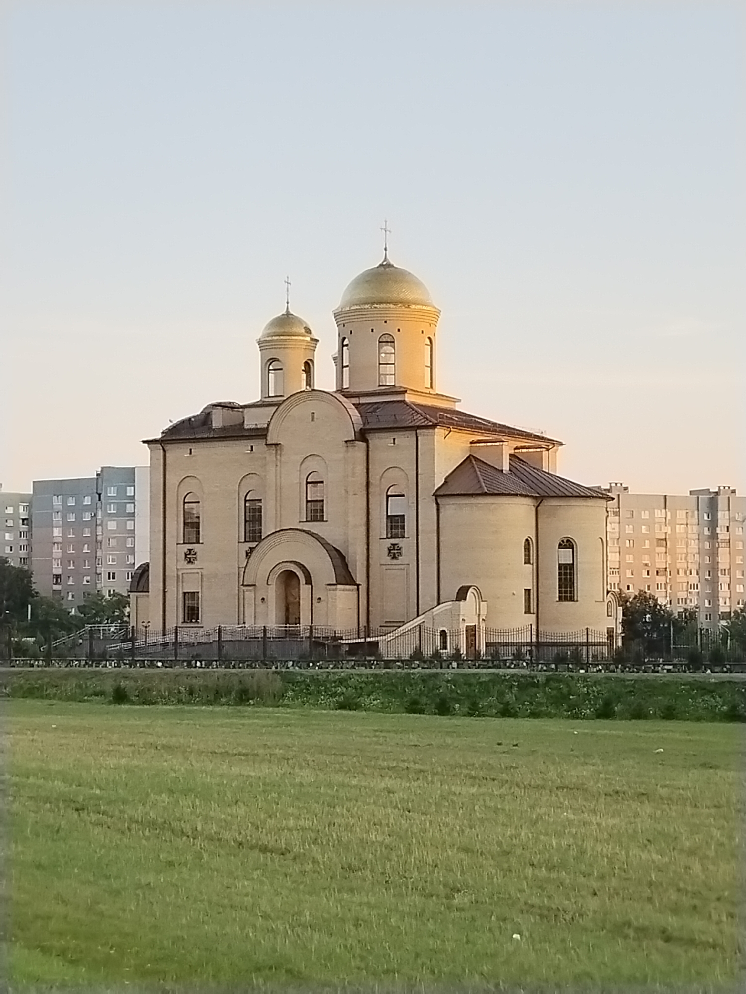 The image captures a large, light-colored church with golden domes, set against a backdrop of apartment buildings. The church features arched windows and a prominent entrance with a curved archway. A black metal fence encloses the church grounds, which appear to be elevated from the surrounding area. In the foreground, a vast expanse of green grass stretches out, suggesting an open field or park. The sky is a pale, clear blue, indicating either early morning or late afternoon. The presence of residential buildings indicates an urban or suburban setting. Specifically, this is the Holy Intercession Cathedral located in Hrodna, Belarus. No people are visible in the frame.