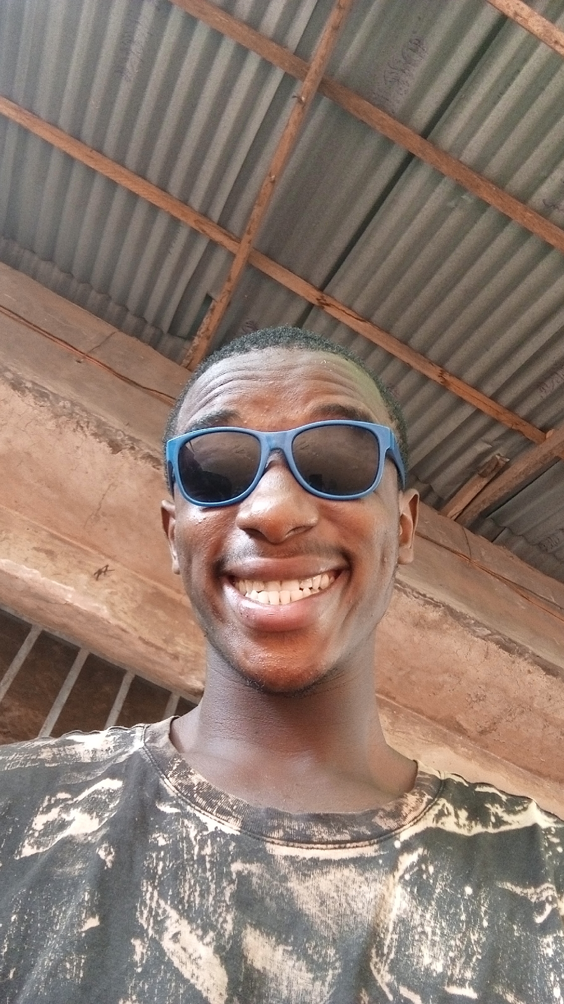 A close-up, medium-shot photograph shows a dark-skinned man smiling broadly while wearing blue sunglasses. He is facing the camera. He is wearing a T-shirt with a tie-dye pattern. The background shows the underside of a corrugated metal roof with wooden support beams. The location is in the city of Unknown, Unknown.