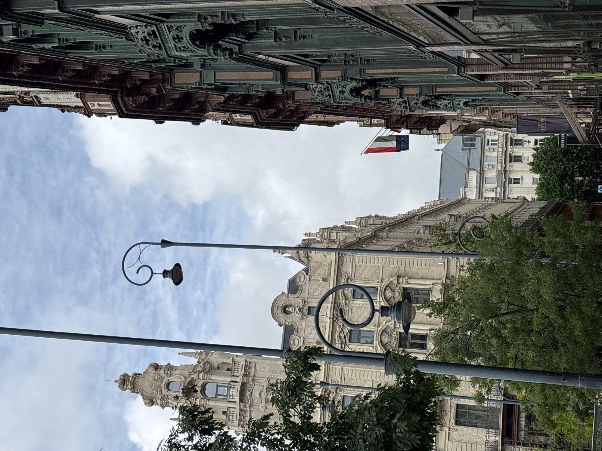 The image shows a street in Budapest, Hungary, taken from a low angle looking upwards. The buildings on either side of the street display elaborate architectural details, including carvings and ornate trim. On the left side of the image, the buildings have a darker, more ornate facade, while the buildings on the right side feature a lighter color palette. Street lamps with decorative coiled arms are visible, along with trees that obscure some of the buildings. A set of flags, including what appears to be the flag of Kuwait, are seen flying between the buildings. The sky above is partly cloudy.