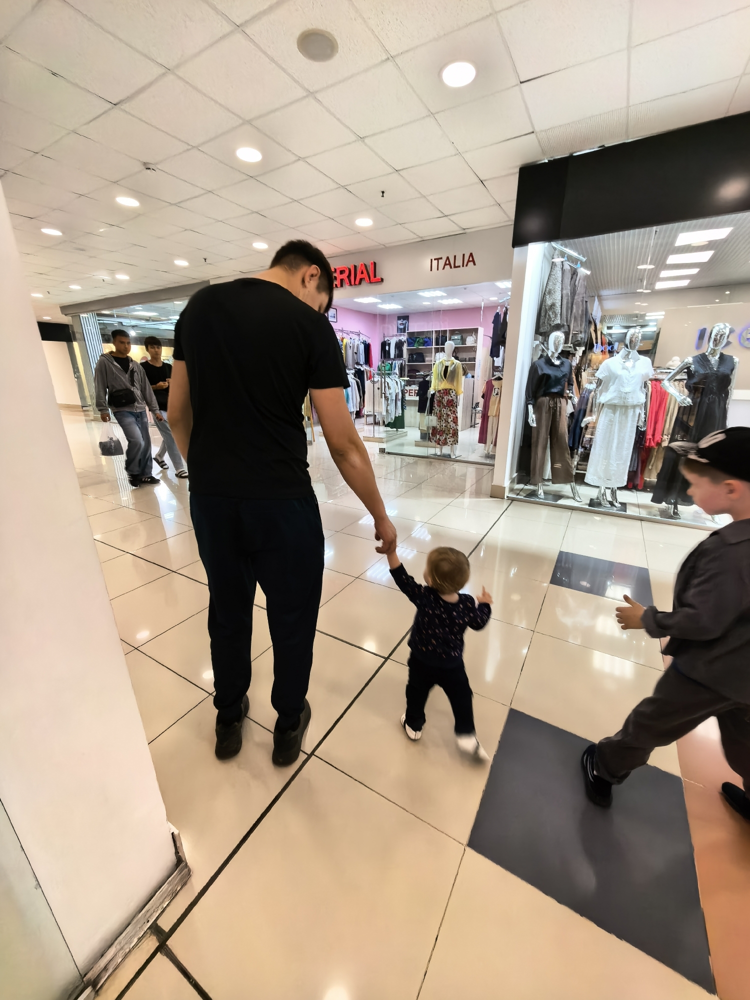 In Sterlitamak, Russia, a family is walking through a mall. A man in a black t-shirt and dark pants holds the hand of a toddler dressed in a dark, patterned outfit. The toddler is taking steps, and a young boy in a grey jacket and dark pants walks alongside them. In the background, two young men walk away from the camera, one wearing a grey hoodie and the other a black t-shirt. The mall features brightly lit ceilings, white tiled floors with dark grout lines, and a store with mannequins displaying clothing. The store signage reads "ITALIA" in red letters.