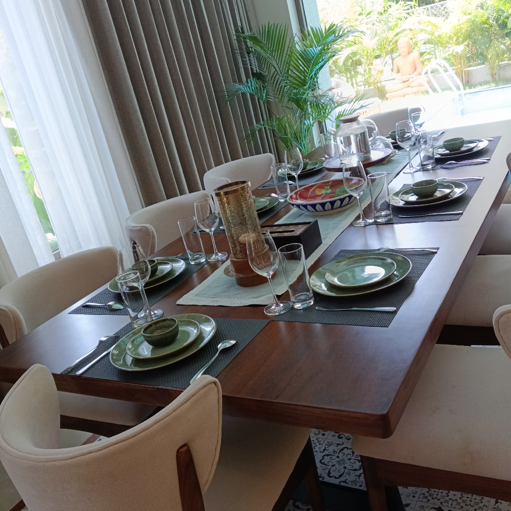 A long dark wood table is set with place settings for eight, each featuring a dark gray placemat, green plates and bowls, and silverware. A light green table runner stretches down the center of the table, adorned with a large decorative bowl, candle holder, and tissue box. Clear glasses and water carafes are also arranged along the runner. The table is surrounded by upholstered chairs with a light beige hue.
A large window with sheer white curtains and heavy gray drapes provides a view to the outdoors, revealing a tropical garden with a swimming pool. A Buddha statue is visible in the background. The setting appears to be in a private home, likely located in Ashvem, India, based on the lush tropical vegetation and architectural style. No people are visible in the image.