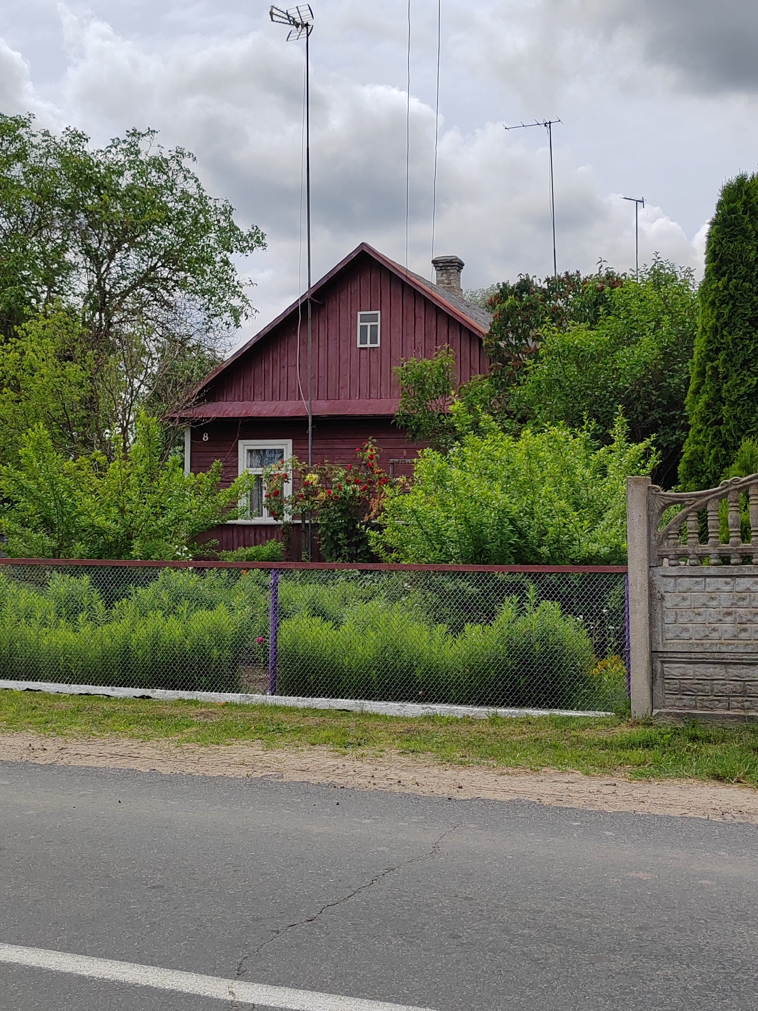 A view of a detached, dark red wooden house with a pitched roof, located in Воўчынскі сельскі Савет, Belarus. The house has a small, square window on the upper portion of its gable end and a larger window on the side. A white-framed window with visible panes is located on the main body of the house. Adjacent to the house is a fence made of wire mesh with a dark metal top frame and a lighter grey stone gatepost on the right. The area in front of the house and fence is overgrown with green bushes and some flowering plants. In the background, several tall, thin antennas are visible against a cloudy sky. A paved road with a white stripe runs in the foreground of the image.