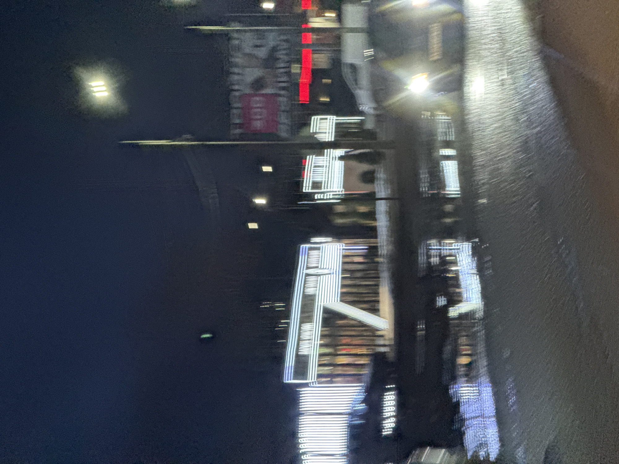 At night, the image shows a street in Chaikovsky, Russia, with building lights reflecting off a wet road surface. Various illuminated signs are visible, with one sign displaying the name "CHAIKOVSKY" in vertical lettering. There are also streetlights and indications of vehicular traffic.