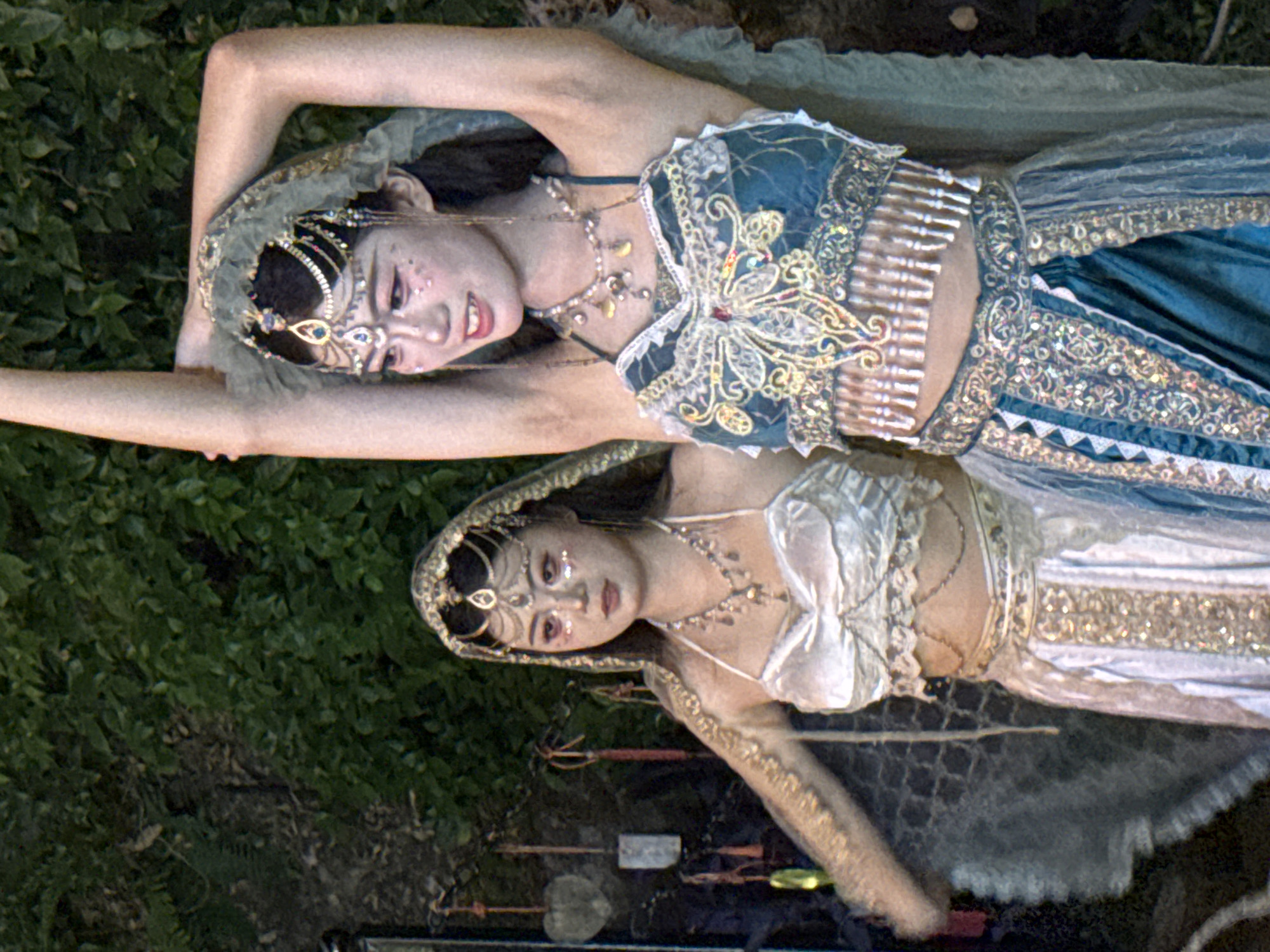 Two young women dressed in elaborate, traditional Indian-inspired costumes with shimmering sequins and gold embroidery are posing outdoors. The woman on the right, facing the viewer, has her arms raised above her head, and her costume is a teal blue halter top with a beaded fringe and a layered skirt. The woman on the left, facing slightly away from the viewer, wears a white halter top and a white skirt with gold trim. Both women have ornate headpieces with chains and jewels, and their makeup includes glitter on their cheeks and eyes. They are positioned in front of a dark, leafy green background that appears to be foliage. The overall scene suggests a performance or a festive event. There are some hanging decorations to the left of the woman on the left, including what appears to be a carved wooden heart and some red and yellow tassels. The image does not contain any specific elements that would indicate a location within Yuzhong District, China.