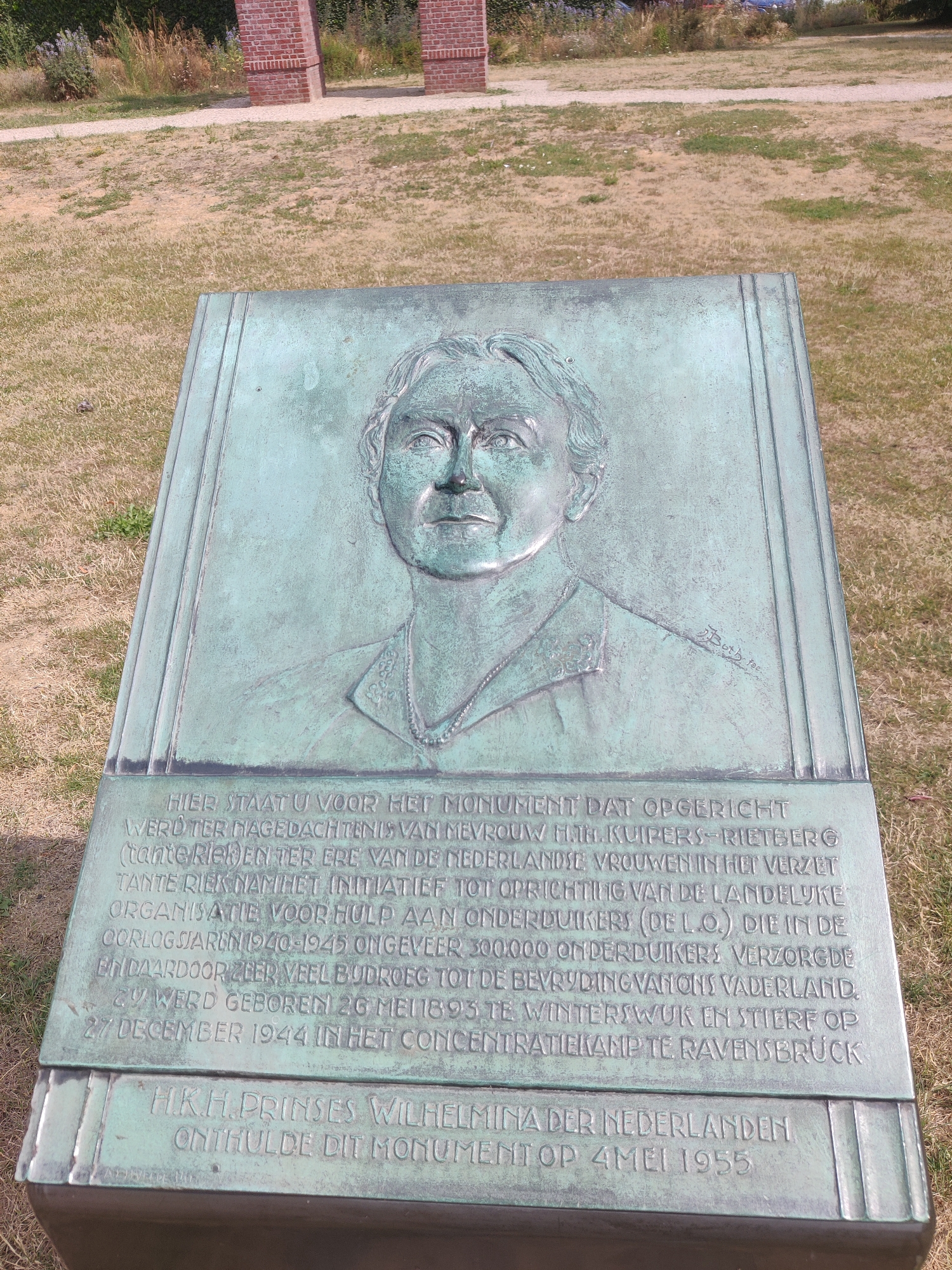 The image shows a bronze relief plaque with an inscription, mounted on a stone base. The plaque depicts a portrait of a woman in profile, with a necklace and collared jacket. The inscription below the portrait is in Dutch and provides details about the monument. The monument is located in Winterswijk, Netherlands. The inscription reads: "HIER STAAT U VOOR HET MONUMENT DAT OPGERICHT WERD TER NAGEDACHTENIS VAN MEVROUW H.H. KUIPERS-RIETBERG (tante Riet) EN TER ERE VAN DE NEDERLANDSE VROUWEN IN HET VERZET. TANTE RIET NAM HET INITIAF TOT DE LANDELIJKE ORGANISATIE VOOR HULP AAN ONDERDUIKERS (DE L.O.). DIE IN DE OORLOGSJAREN 1940-1945 ONGEVEER 300.000 ONDERDUIKERS VERZORGDE EN DAARDOOR ZEER VEEL BIJDROEG TOT DE BEVRIJDING VAN ONS VADERLAND. ZIJ WERD GEBOREN 2G MEI 1895 TE WINTERSWIJK EN STIERF OP 27 DECEMBER 1944 IN HET CONCENTRATIEKAMP TE RAVENSBRÜCK." Below this inscription, there is a further inscription which reads: "H.K.H. PRINSES WILHELMINA DER NEDERLANDEN ONTHULDE DIT MONUMENT OP 4 MEI 1955". The monument is situated outdoors, with a grassy area and a gravel path visible in the background. There are also some brick structures in the distance. The overall scene suggests a memorial site.