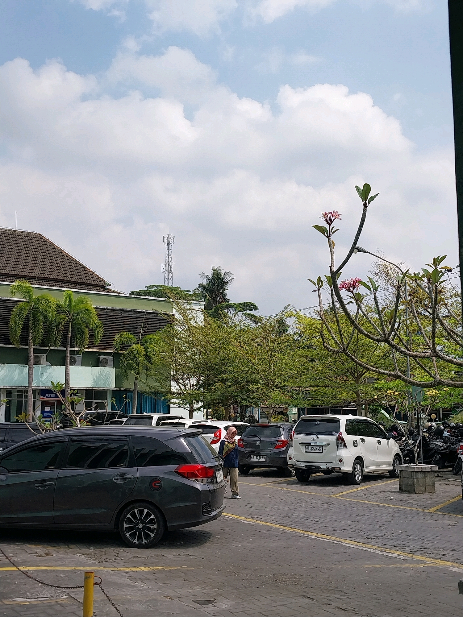The image shows a parking lot outside a building in Wates, Indonesia. The building has a green facade and a brown tiled roof. There are several cars parked in the lot, including a dark gray hatchback and a white minivan. A few people are visible, one walking and another standing near the cars. In the background, trees and a cell phone tower can be seen. The sky is partly cloudy.